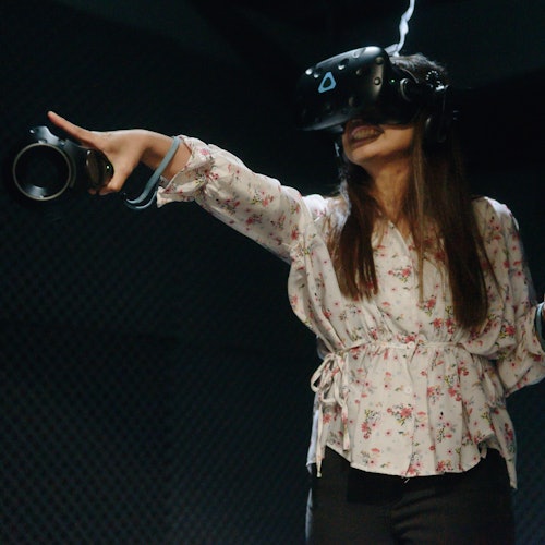 A woman wearing a virtual reality headset and holding controllers points excitedly while engaging with VR content.