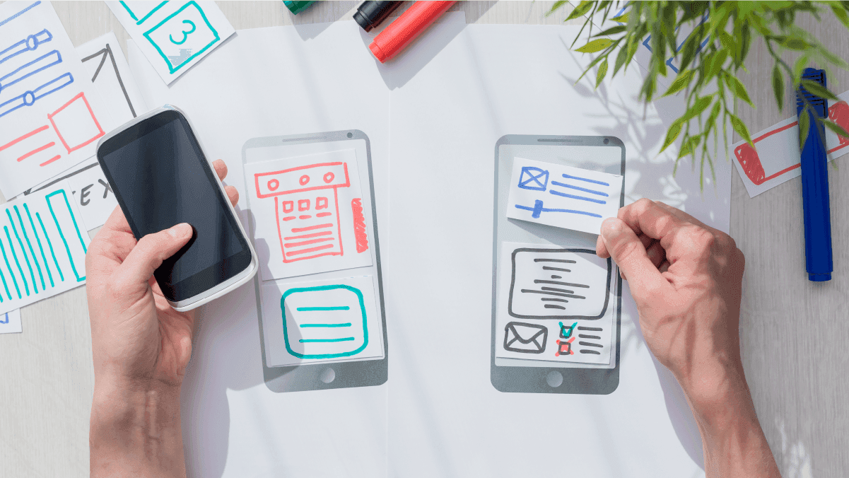 Hands holding a phone over hand-drawn paper interface wireframes of mobile app screens for prototyping