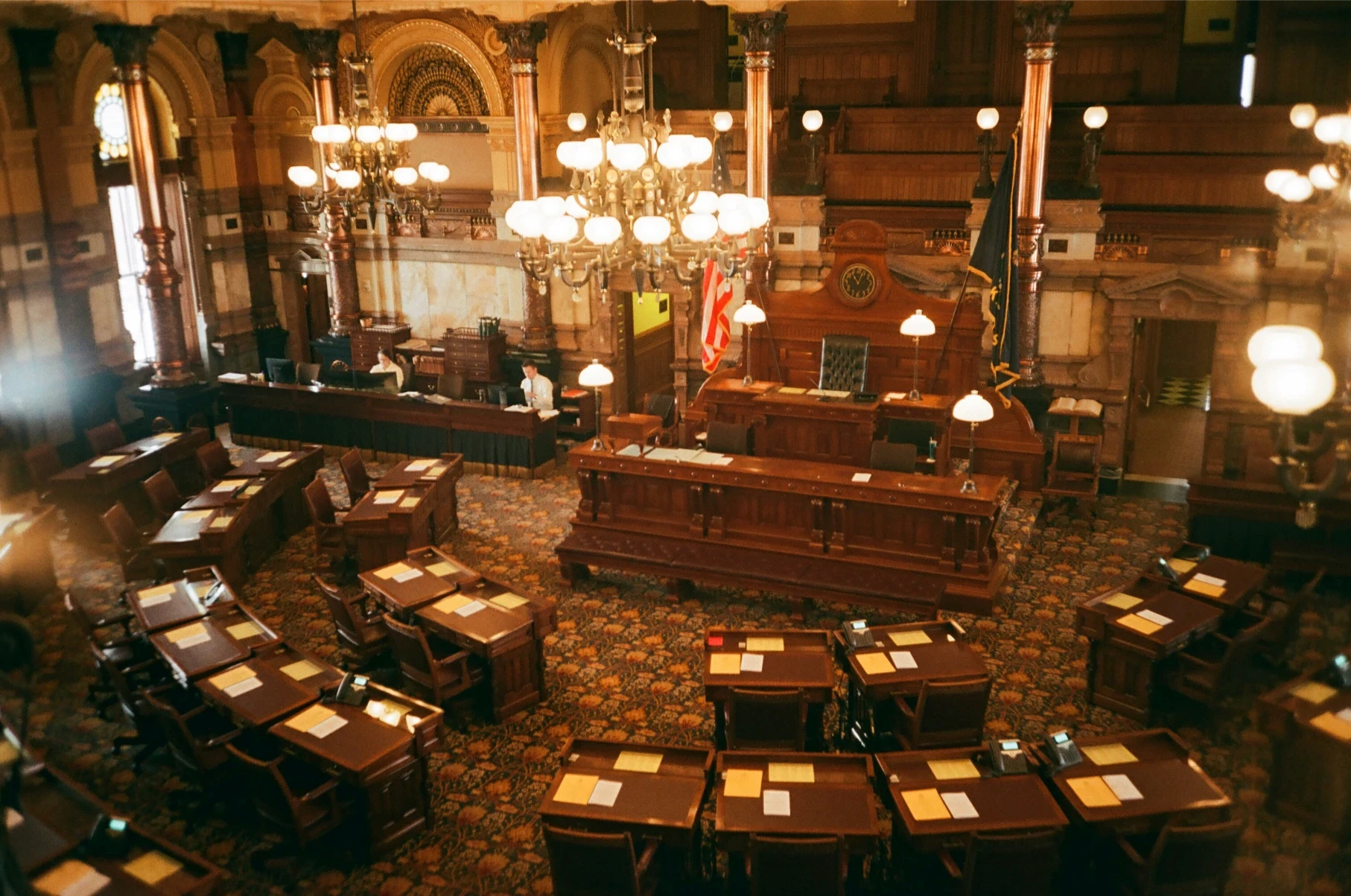 A grand, ornate legislative chamber with wooden desks and chairs arranged in rows. Chandeliers hang from the ceiling, creating a formal and historic ambiance.