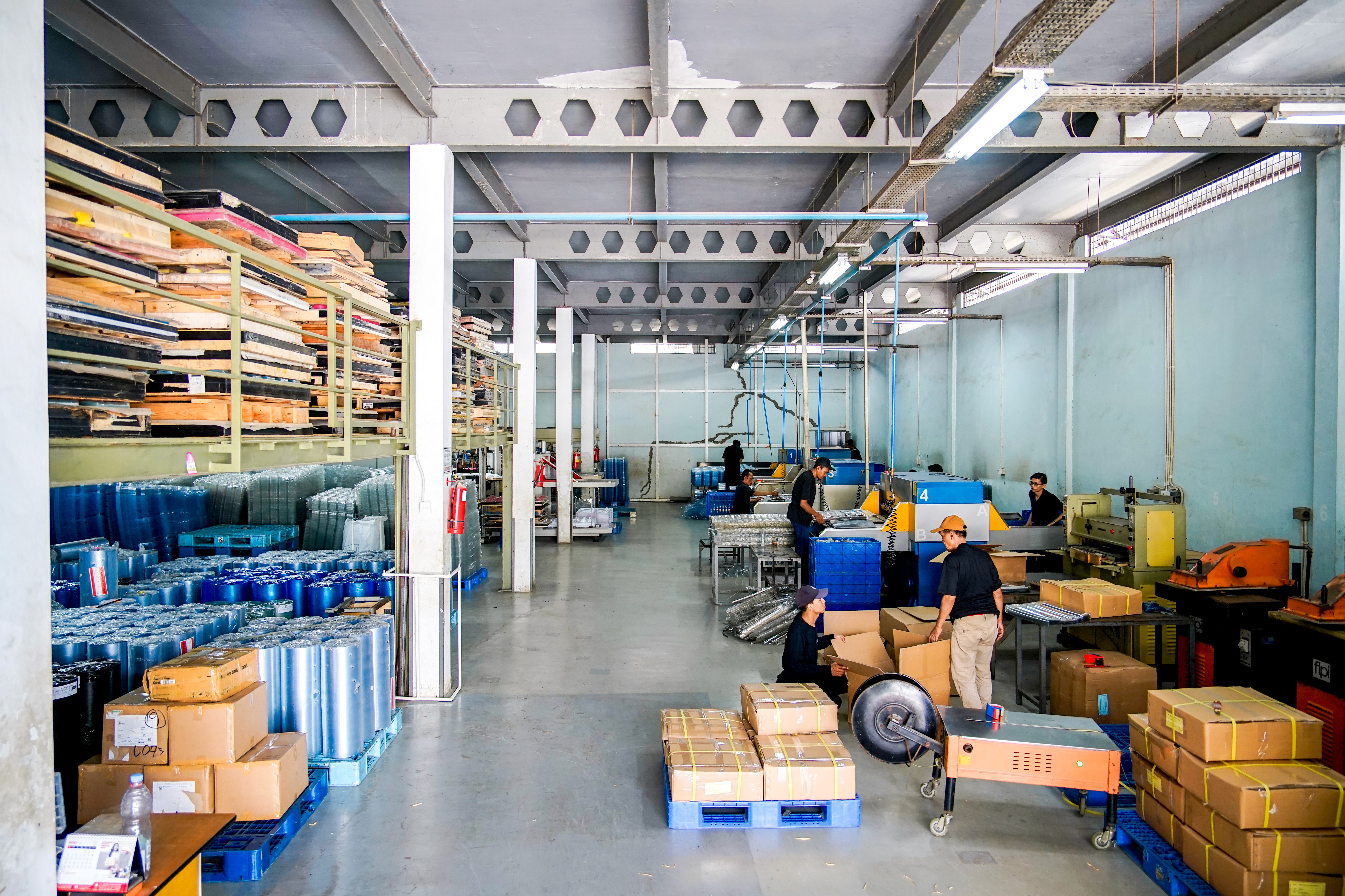 Proses pengepakan dan penyusunan kemasan plastik di gudang (warehouse) sebelum dikirim ke konsumen, terlihat pekerja menyusun produk di atas palet siap kirim dengan latar belakang rak penyimpanan.