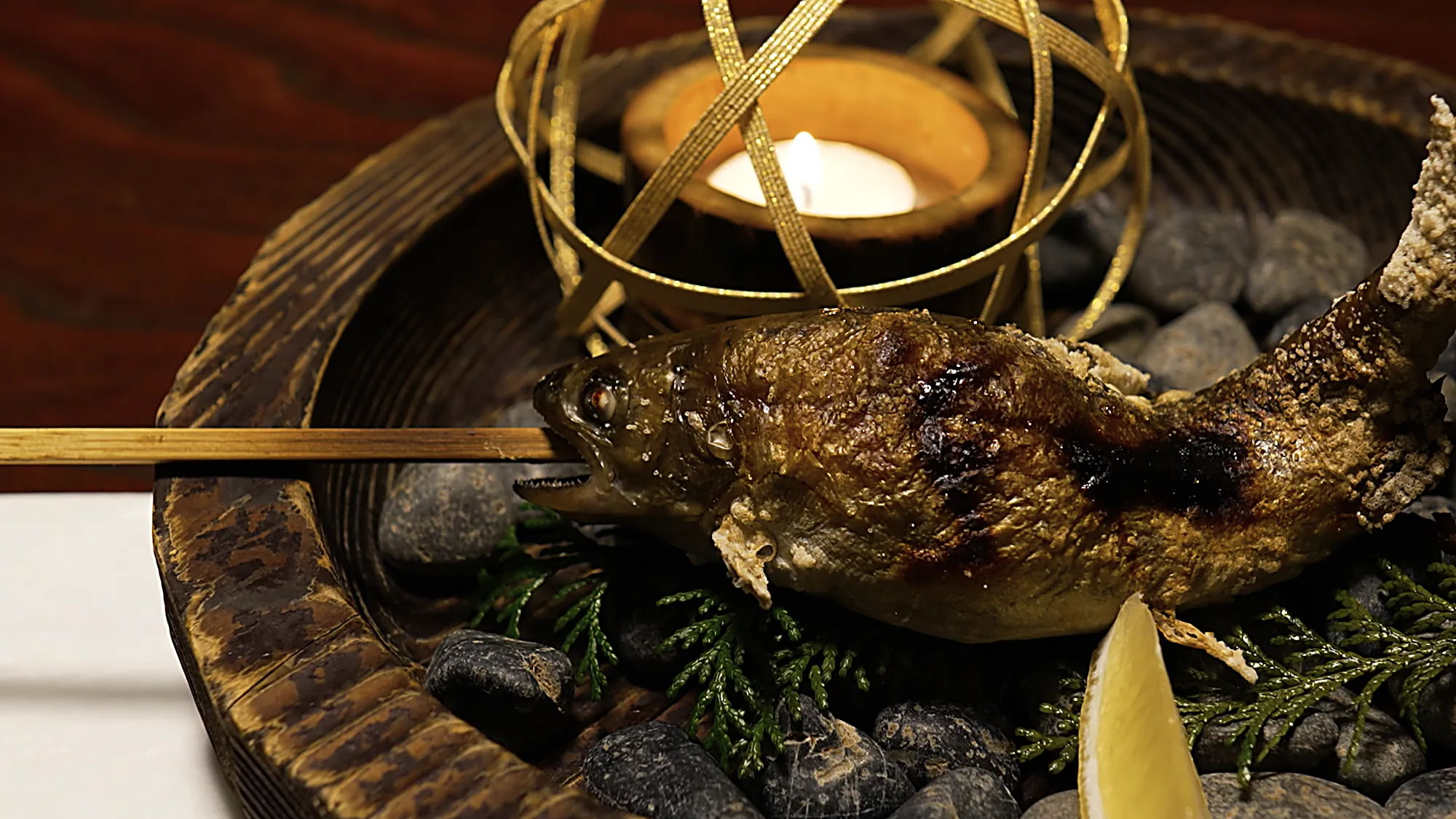 A whole grilled fish on a skewer over stones beside a candle at Kurokawa Onsen
