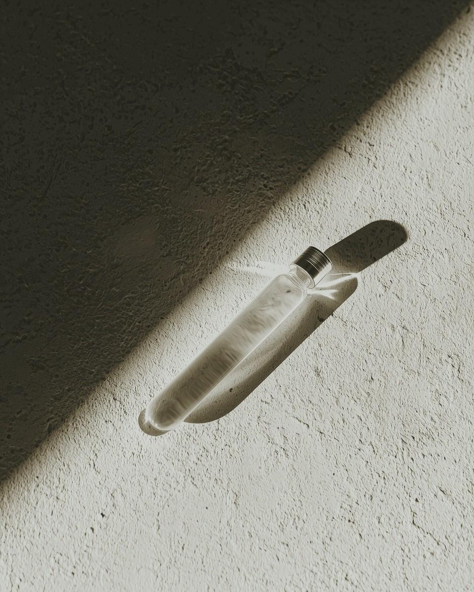 A transparent glass bottle lies on a textured concrete surface, casting a long shadow in the sunlight, creating a minimalist and artistic composition.