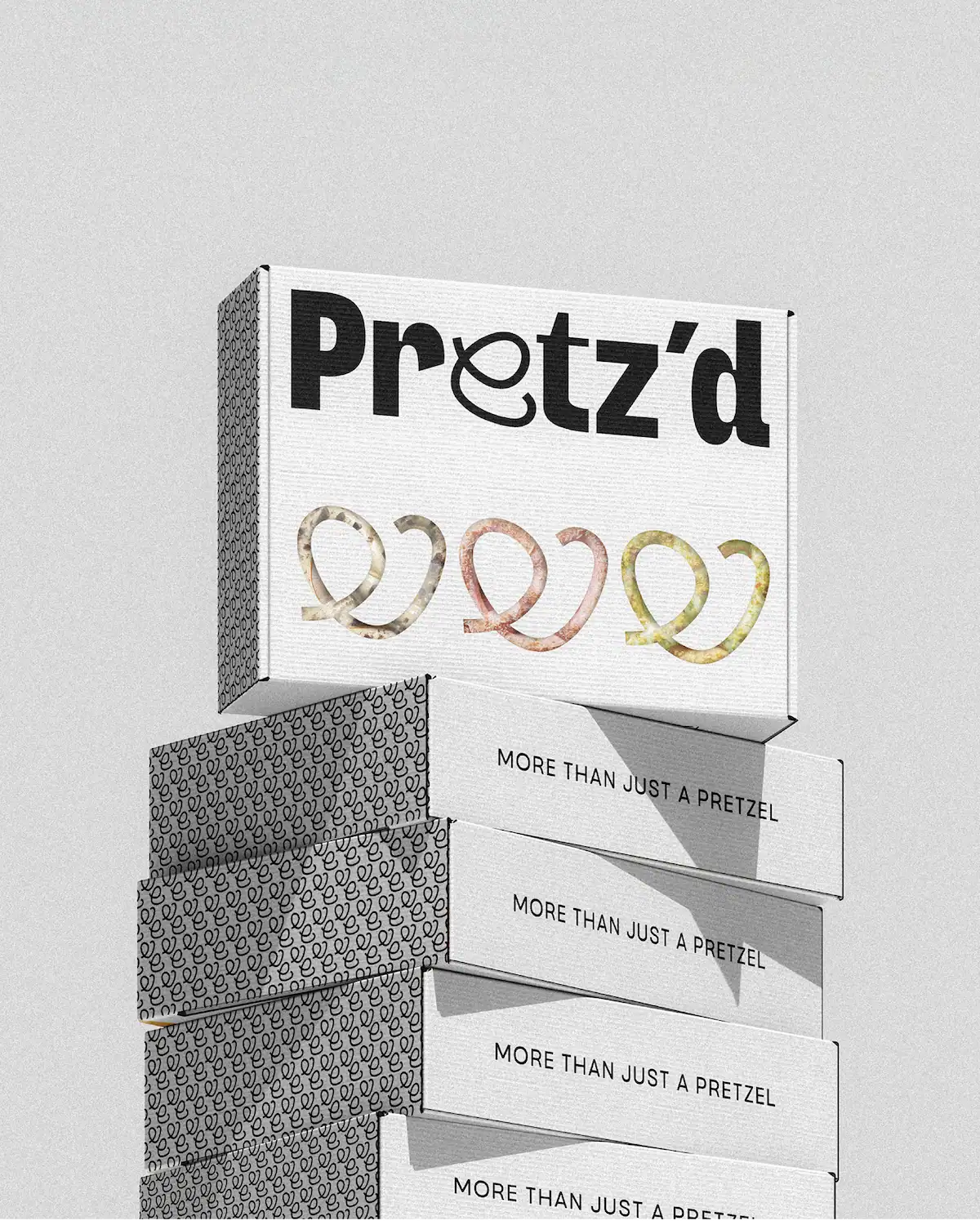 Tour de boîtes d'emballage Pretz'd empilées, face principale avec logo et pictos bretzels colorés, faces latérales avec pattern monochrome et slogan "More than just a pretzel".