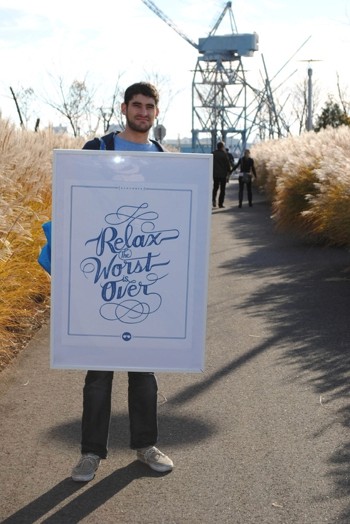 A photo of a man sitting on an alley near a port while holding a white placard with blue handwrite that says: Relax The Worst Is Over.