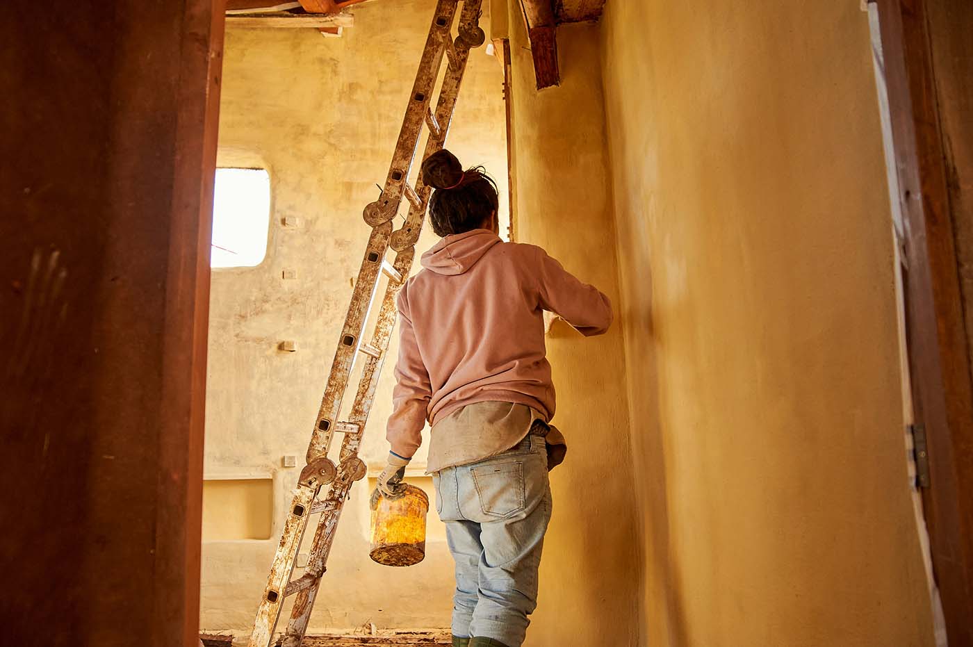 A person wearing a pink hoodie and jeans applying a yellow substance to a wall.
