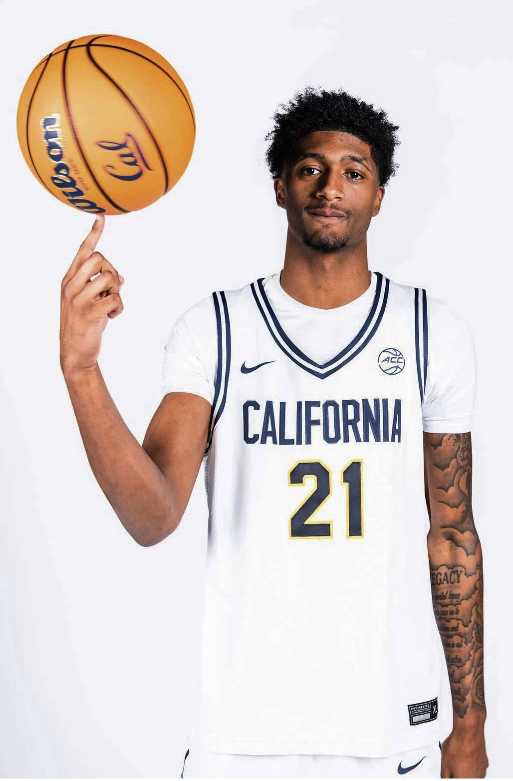 Jake Wilkins wearing a white California jersey with the number 21 spins a basketball on his finger, posing confidently against a plain white background.