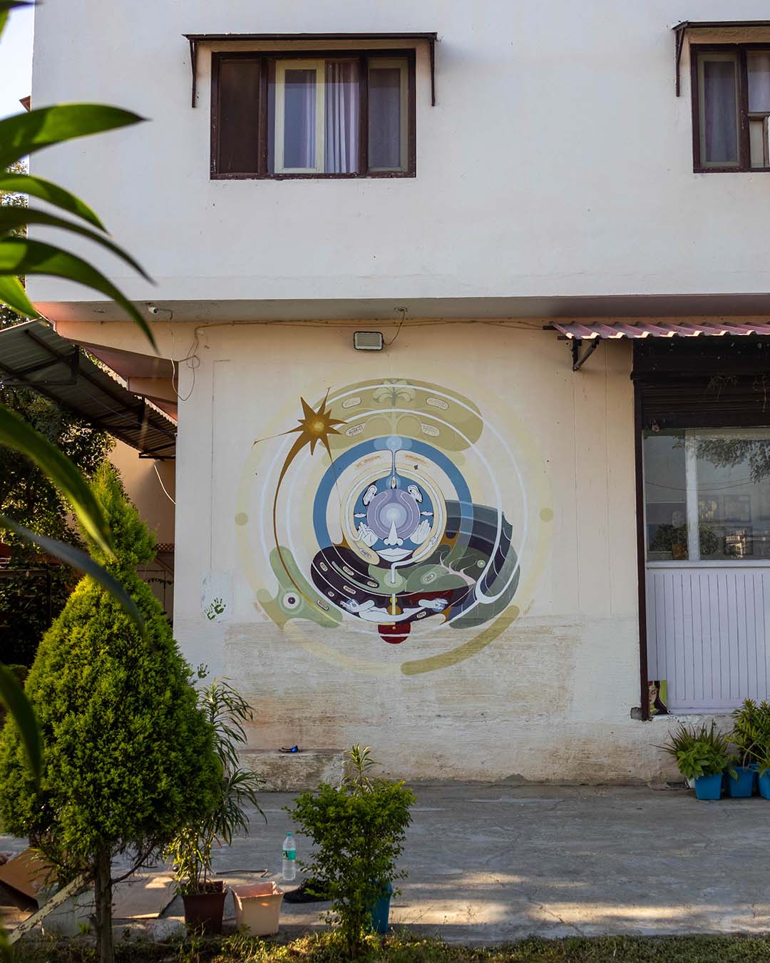 A circular mandala-style mural on a white building near the entrance surrounded by several potted plants next to a garden.