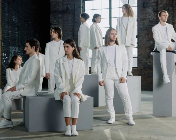 A group of people dressed entirely in white suits are posing on and around gray geometric blocks in a large, industrial space.