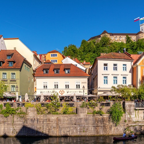 Колоритни рекични сгради с външни места за сядане, буйна зеленина и замък на върха на хълм под ясно синьо небе.