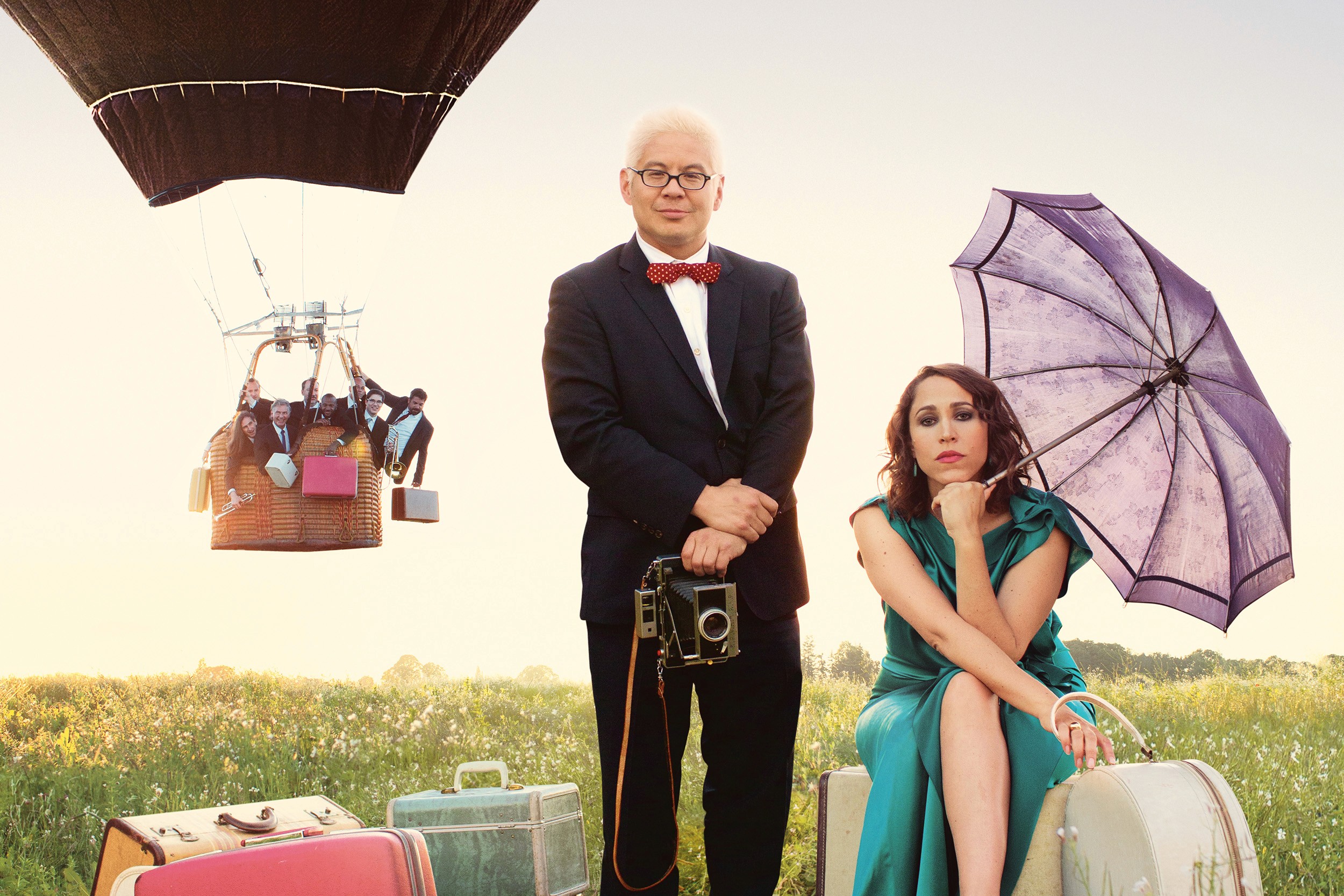 A man with white hair stands next to a woman sitting on suitcases and holding an umbrella in middle of a grass field. In the background, a hot air balloon is rising with bunch of people holding suitcases in the basket.