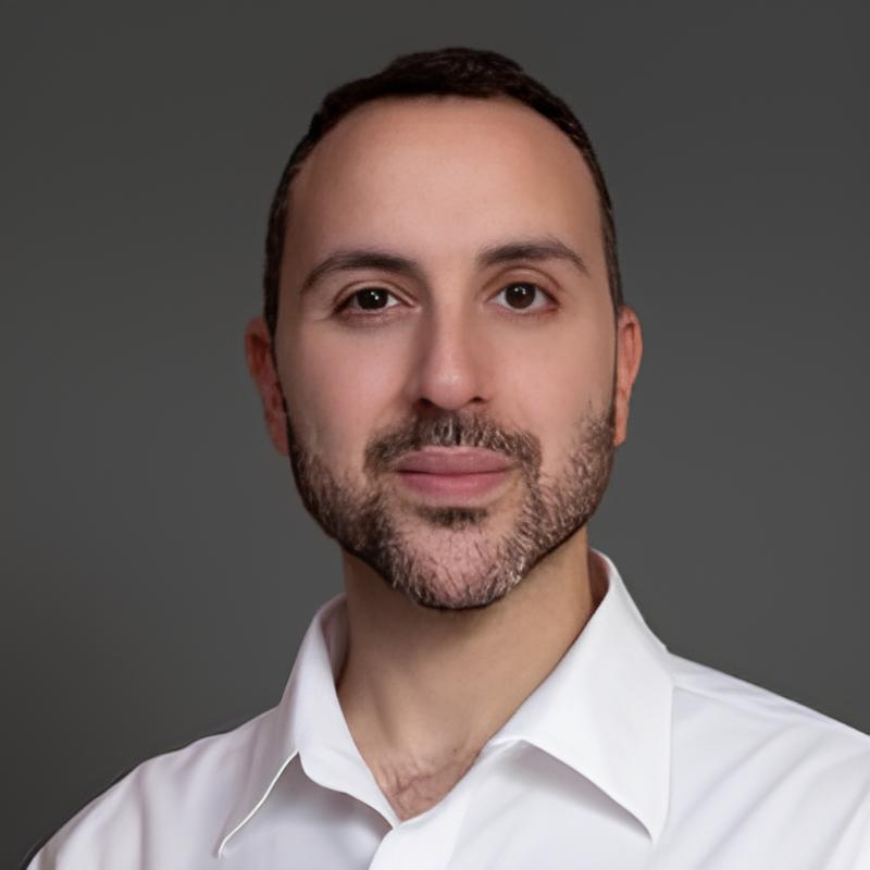 A smiling man with curly hair is sitting at a desk, looking at the camera, with a laptop and a lamp in the background.