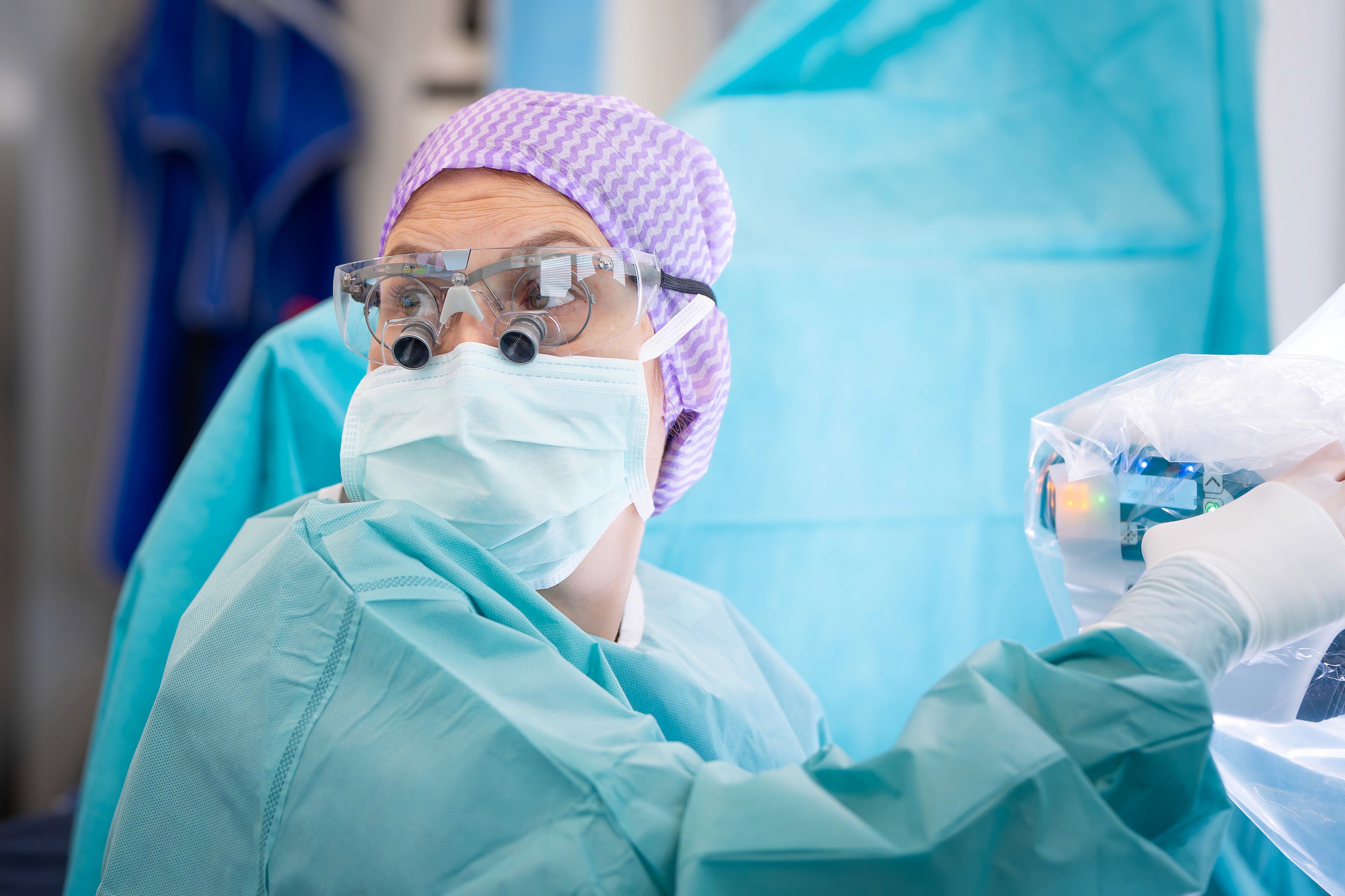 Dr. Christin Kammerhofer mit einer chirurgische Maske, eine Schutzbrille und eine Vergrößerungslupe trägt, blickt in einem Operationssaal zur Seite, umgeben von blauen chirurgischen Tüchern und Geräten.
