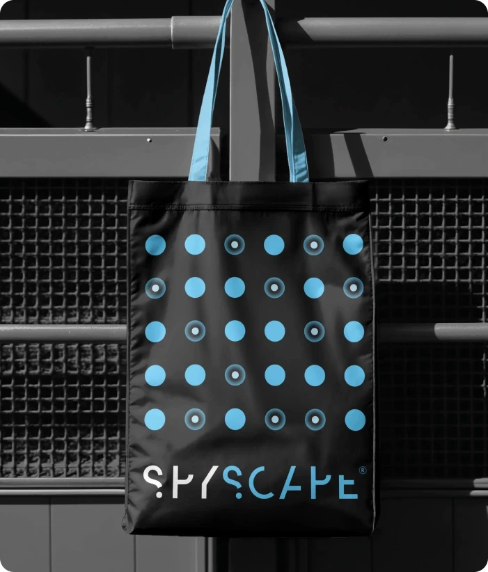 Black SPYSCAPE tote bag with blue dot pattern and SPYSCAPE logo hanging on metal railings.