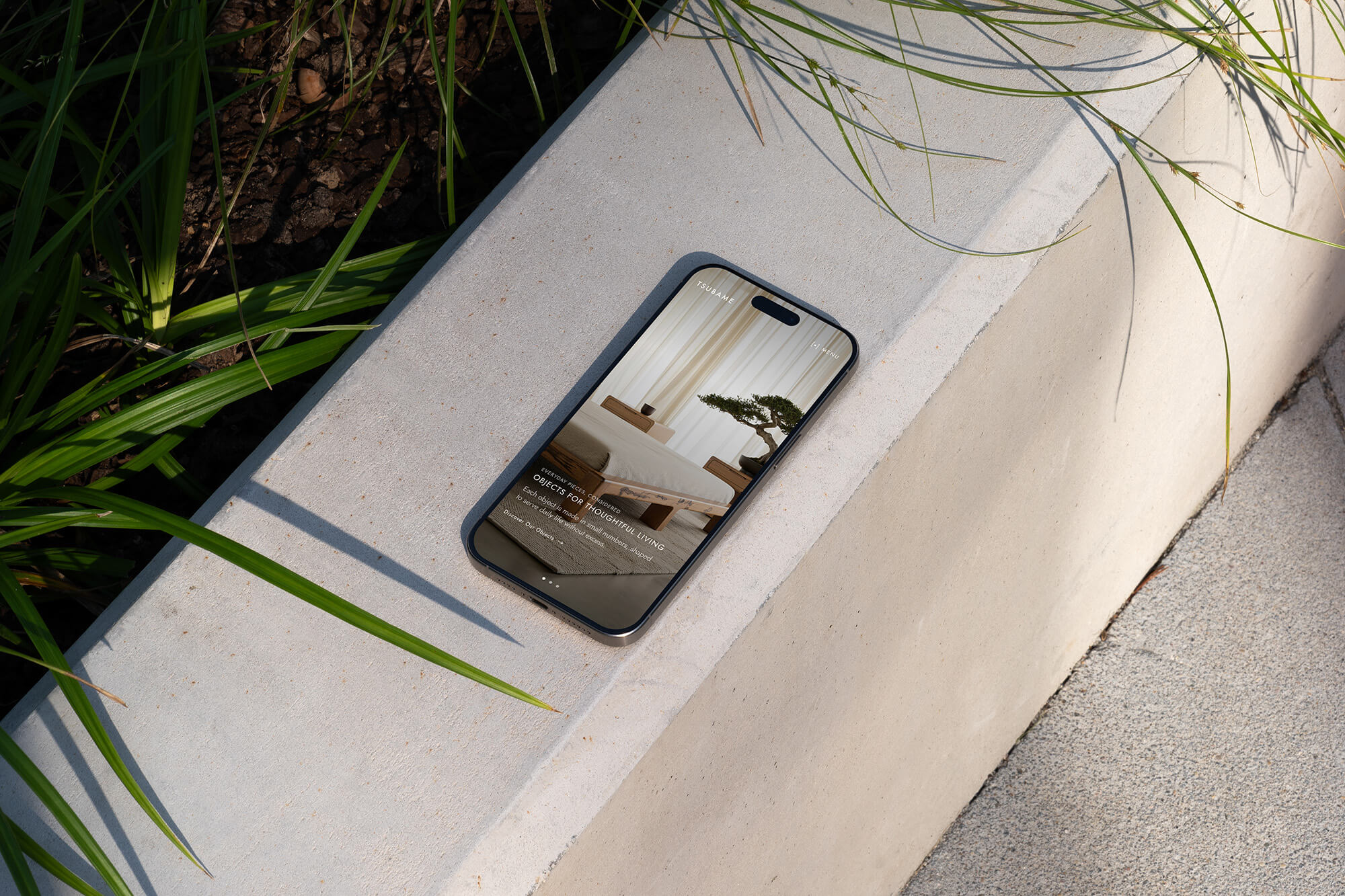 Smartphone on a concrete bench displaying a serene room with a bonsai, surrounded by green leaves, under natural sunlight.