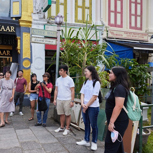A group of seven people stand on a sidewalk near colorful buildings and a shop, with greenery in the background.