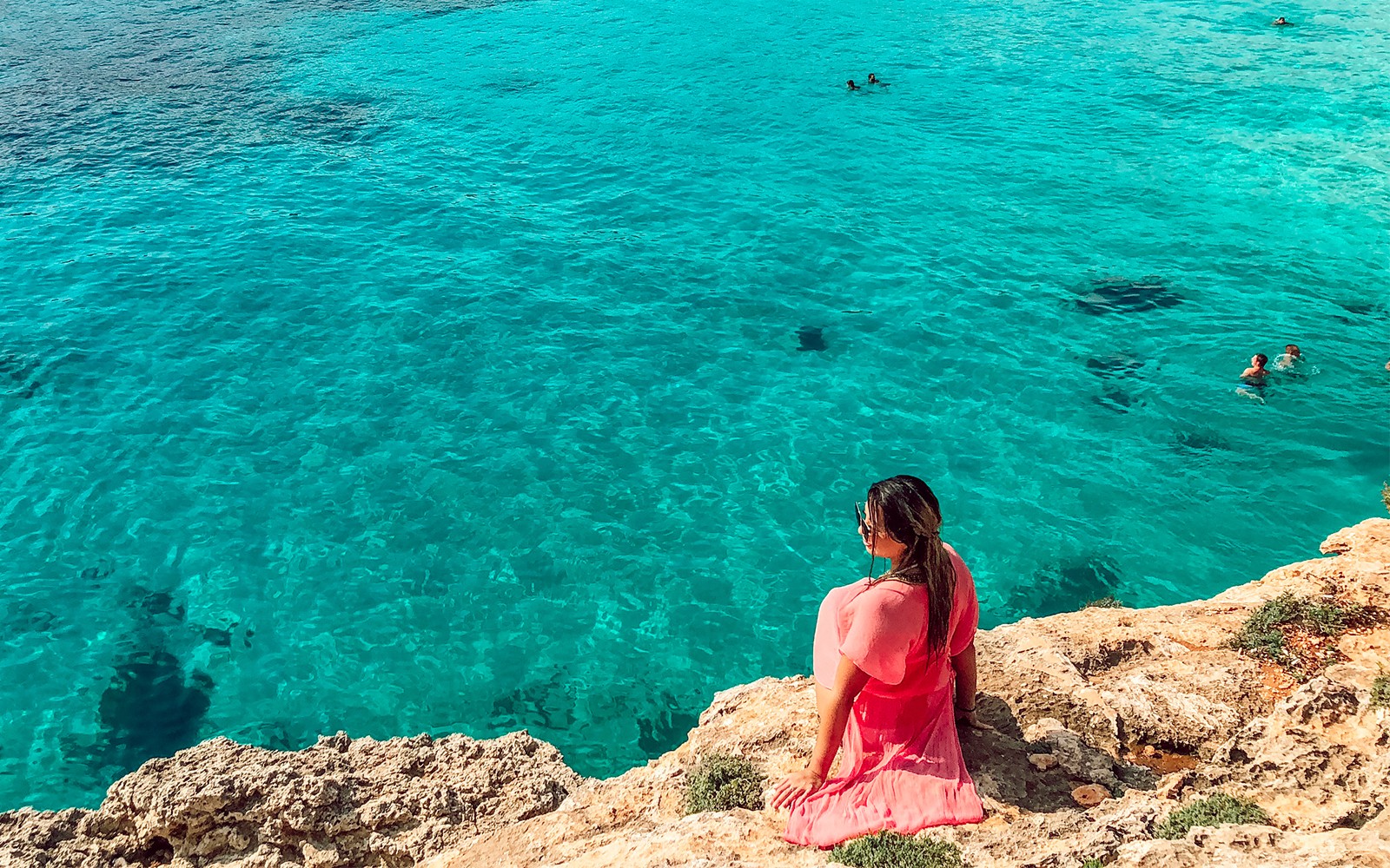 Woman in pink dress sitting on rocky shore overlooking clear blue water in Comino, Malta.