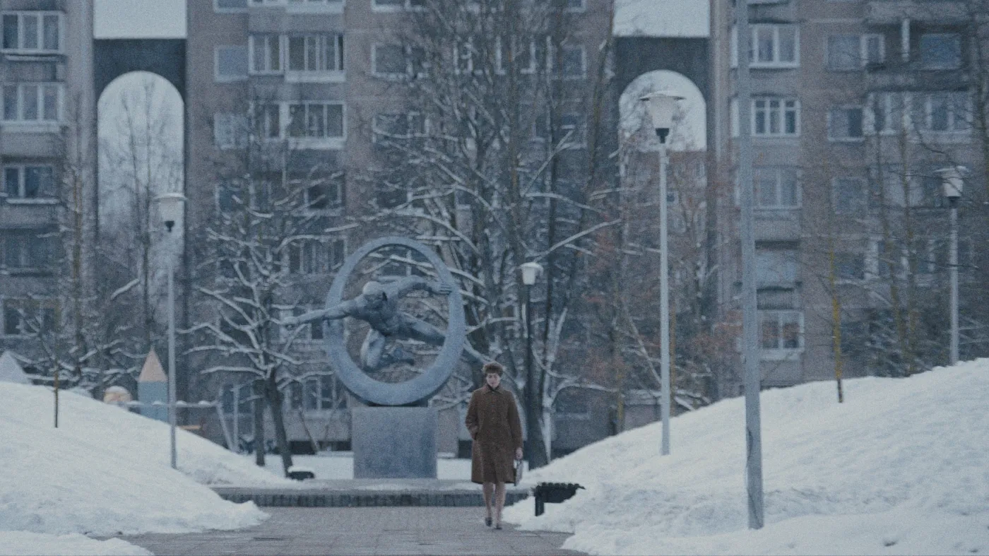 A woman walks through a stark, snow-covered park with leafless trees and a modern sculpture, set against the backdrop of a tall residential building under a cloudy winter sky.