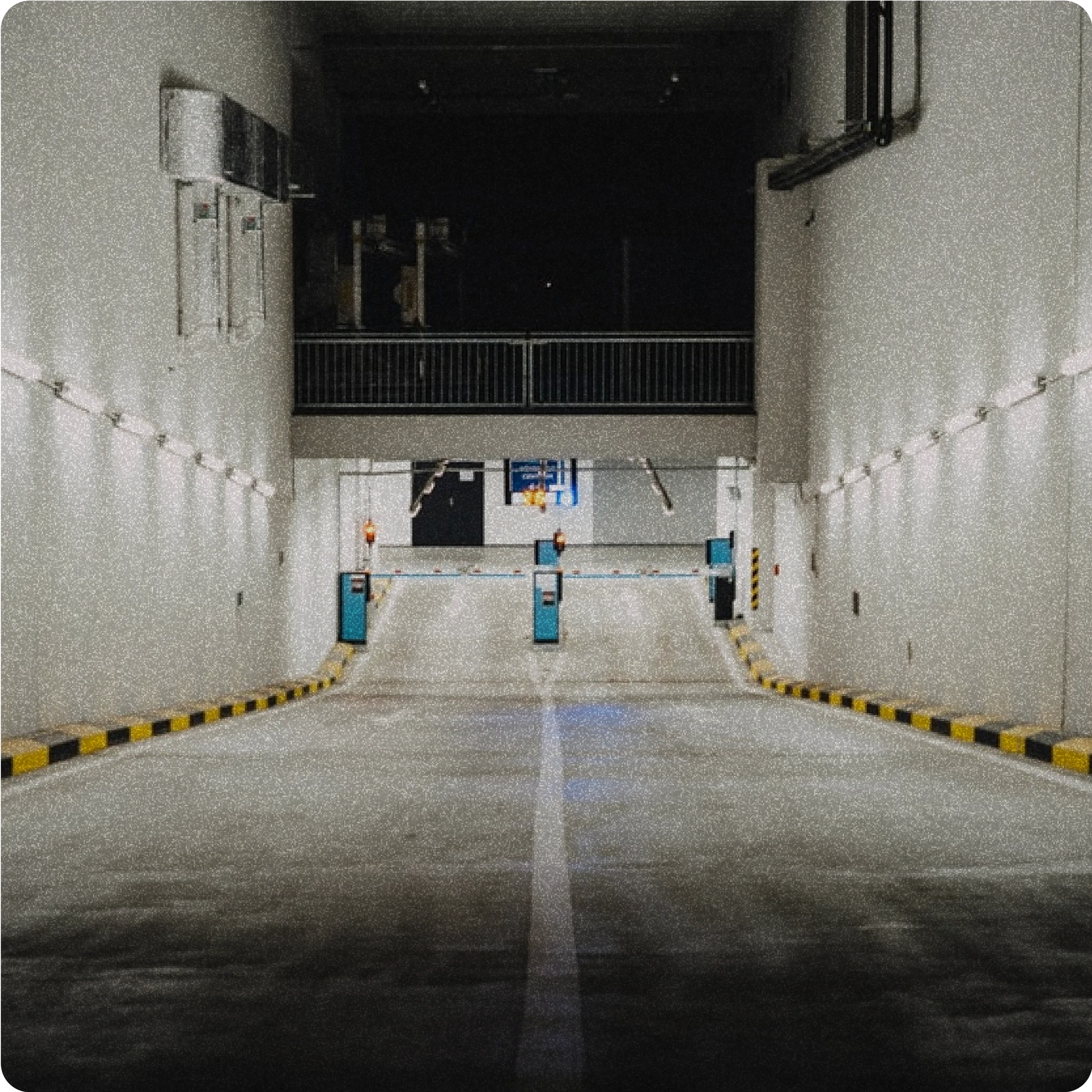 Entrance to an underground parking garage, illuminated by overhead lights, featuring a ramp with barriers and security equipment.