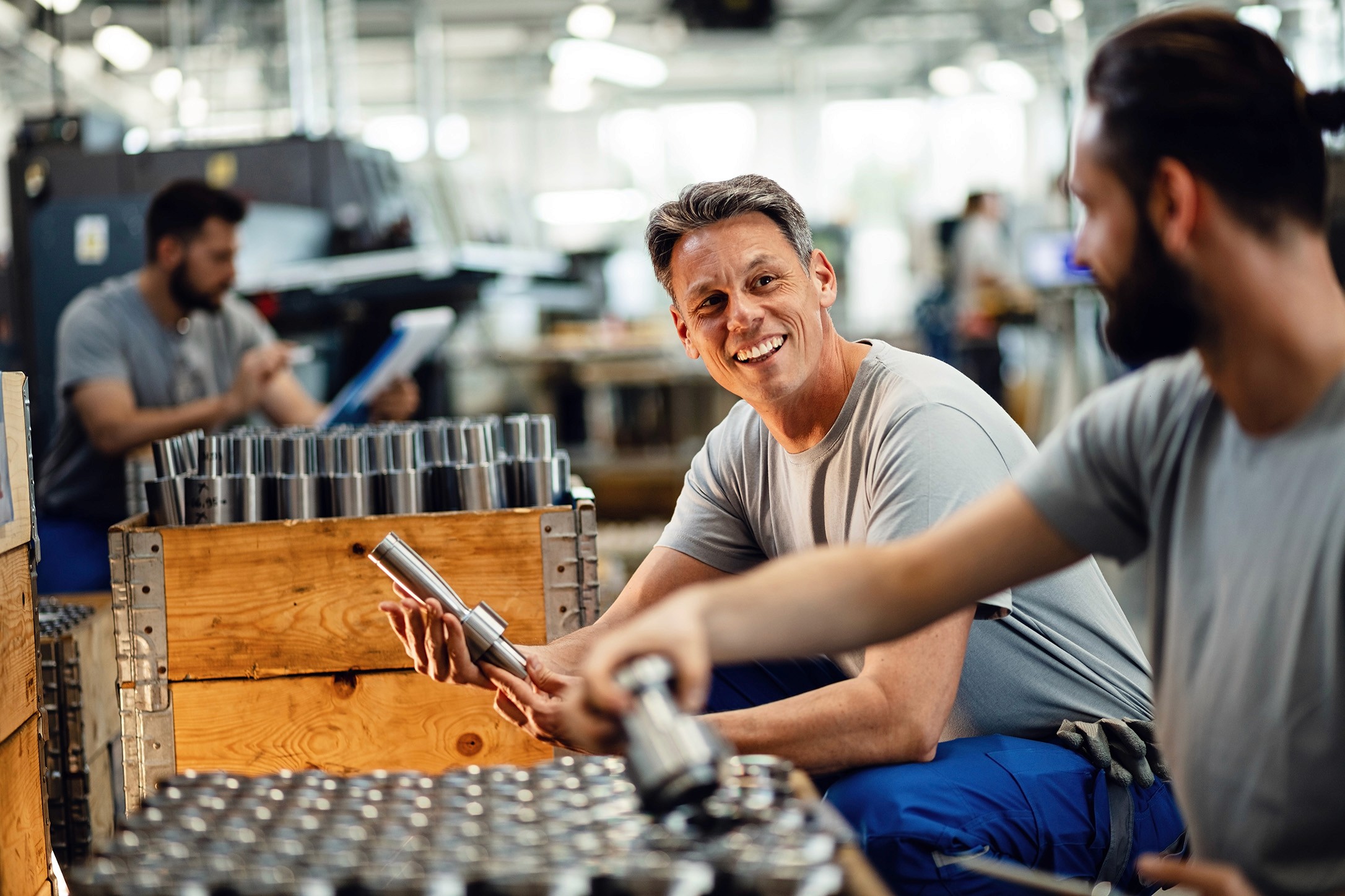 Two smiling male factory workers sorting metal parts. Another worker is in the background, near a machine.