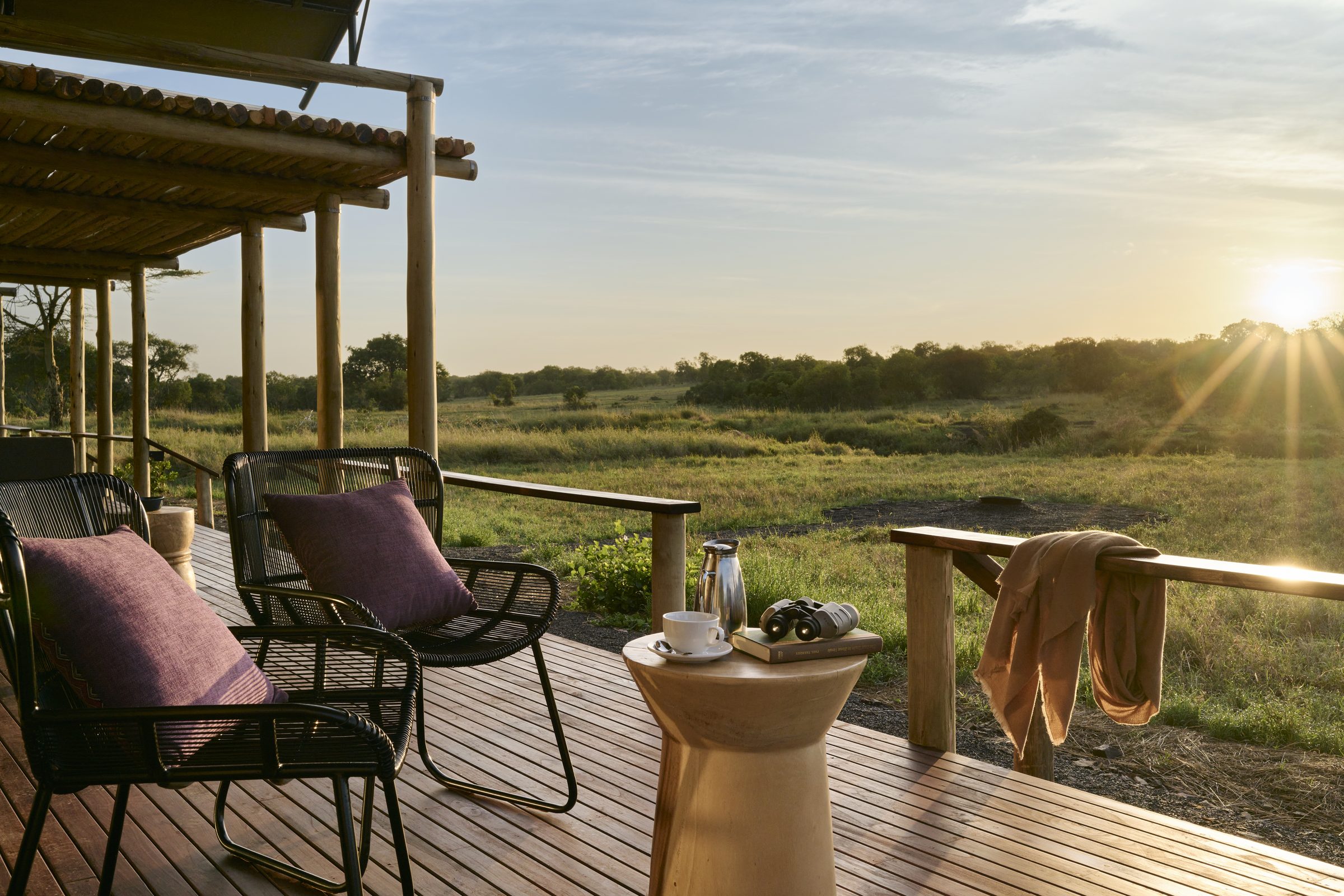 Two cushioned chairs, a side table with coffee and binoculars, and a blanket over the railing look out from a wooden deck across grassland in low sun in Kenya.