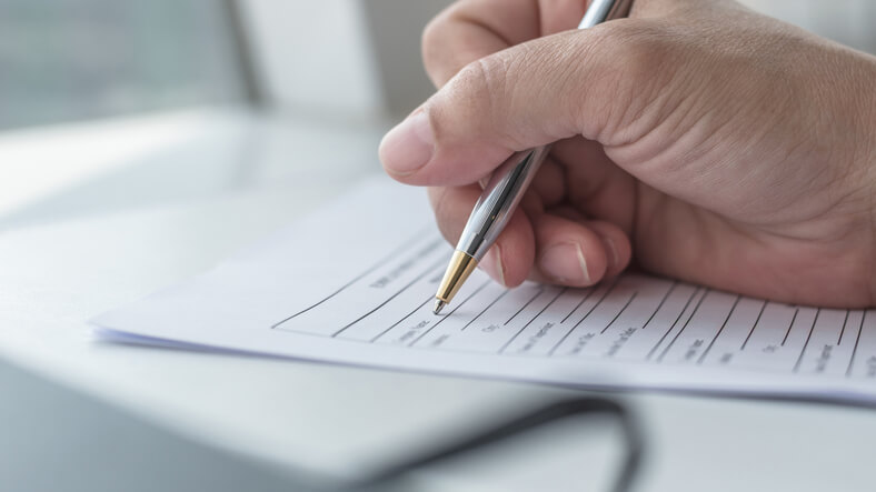 A man's hand holding a pen, writing on a sheet of paper. Form N-600