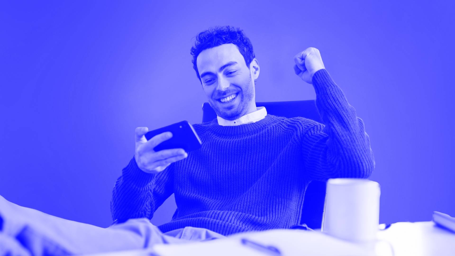 A man smiling with his phone in one hand and his other fist raised with his feet on his desk