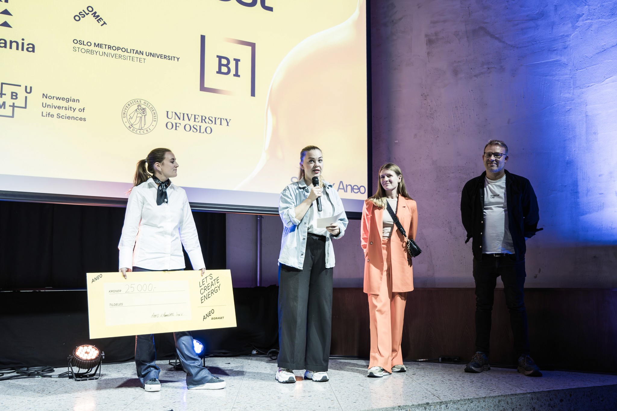 4 people on a stage, including a young woman holding a check to be awarded, while another woman speaks into the microphone. They are at a Spark event where a cash prize is to be awarded to one lucky winner.