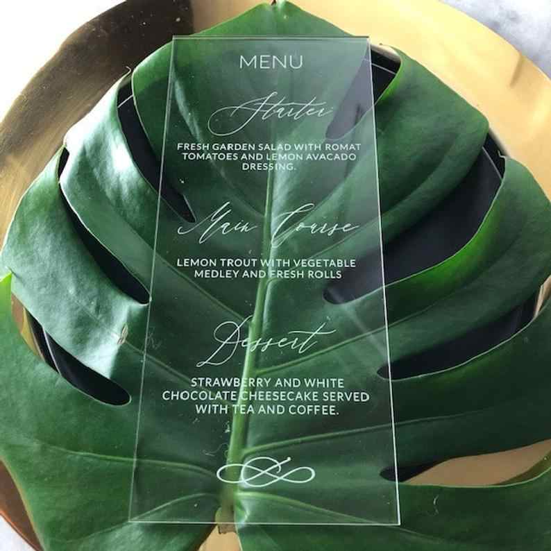 Menu en acrylique transparent avec un texte élégant écrit sur une grande feuille verte posée sur un plateau doré. Il propose des options d'entrée, de plat principal et de dessert.