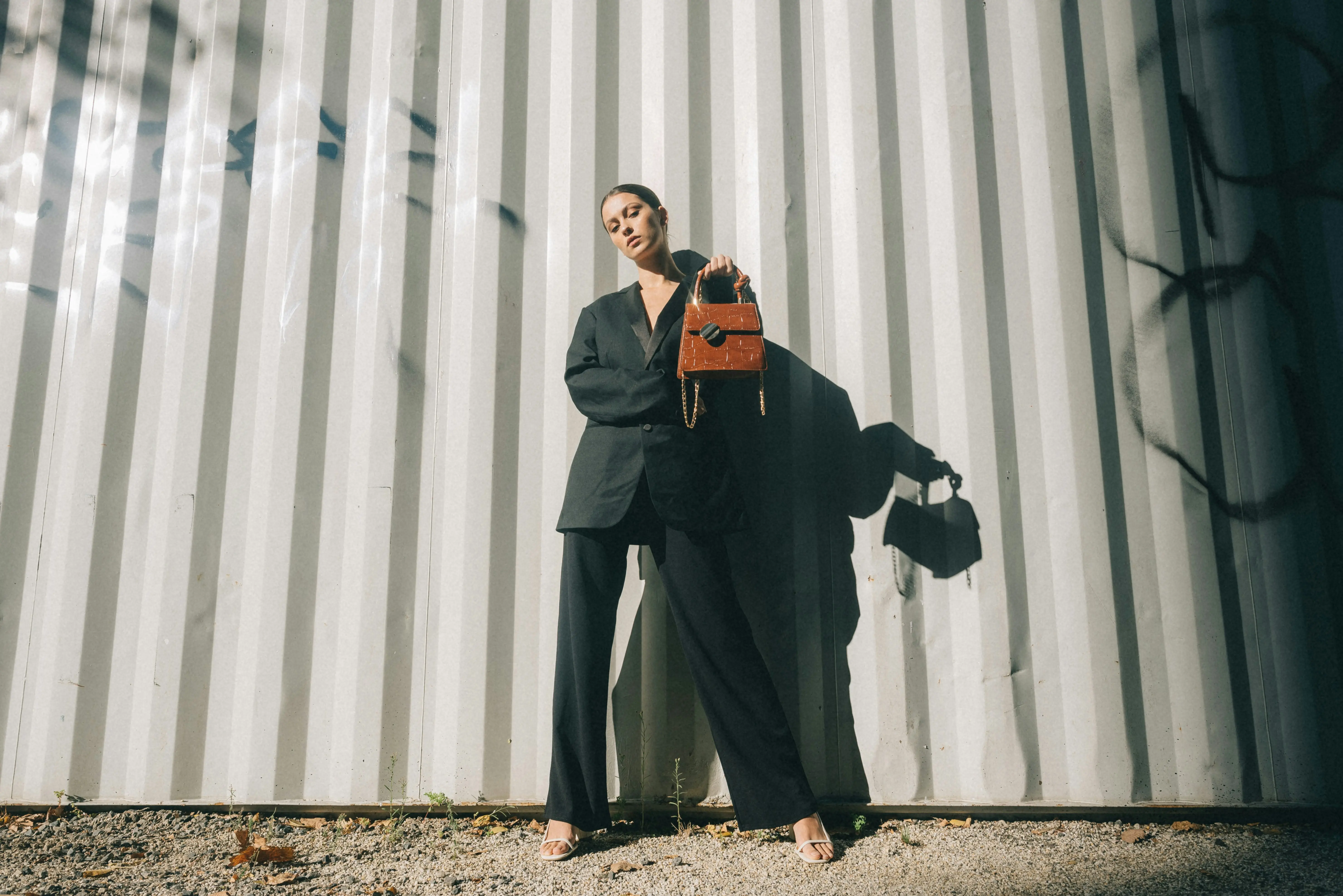 A woman wearing a black blazer and wide-leg trousers stands against a white corrugated metal wall, holding a textured brown handbag.