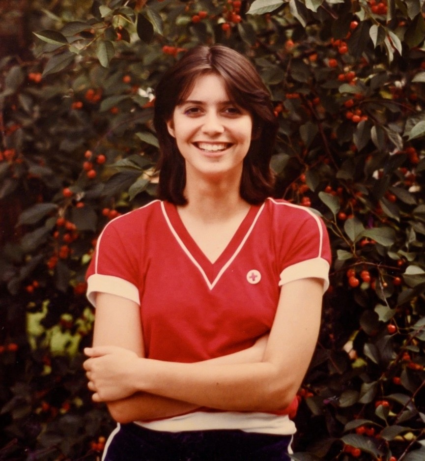 Woman in a red t-shirt standing outside with her arms crossed, smiling.