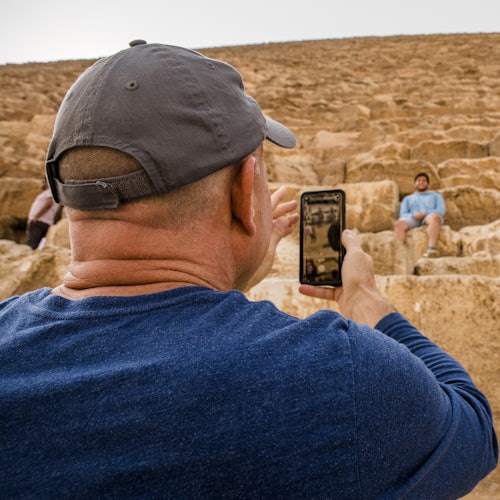 Person wearing a gray cap and blue shirt taking a photo with a smartphone of another person sitting on large stone steps.