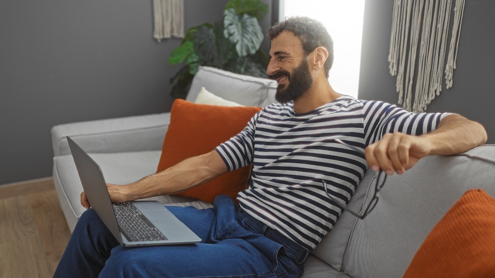 Homme souriant sur un canapé utilisant un ordinateur, explorant les options de renouvellement de son assurance-vie temporaire.