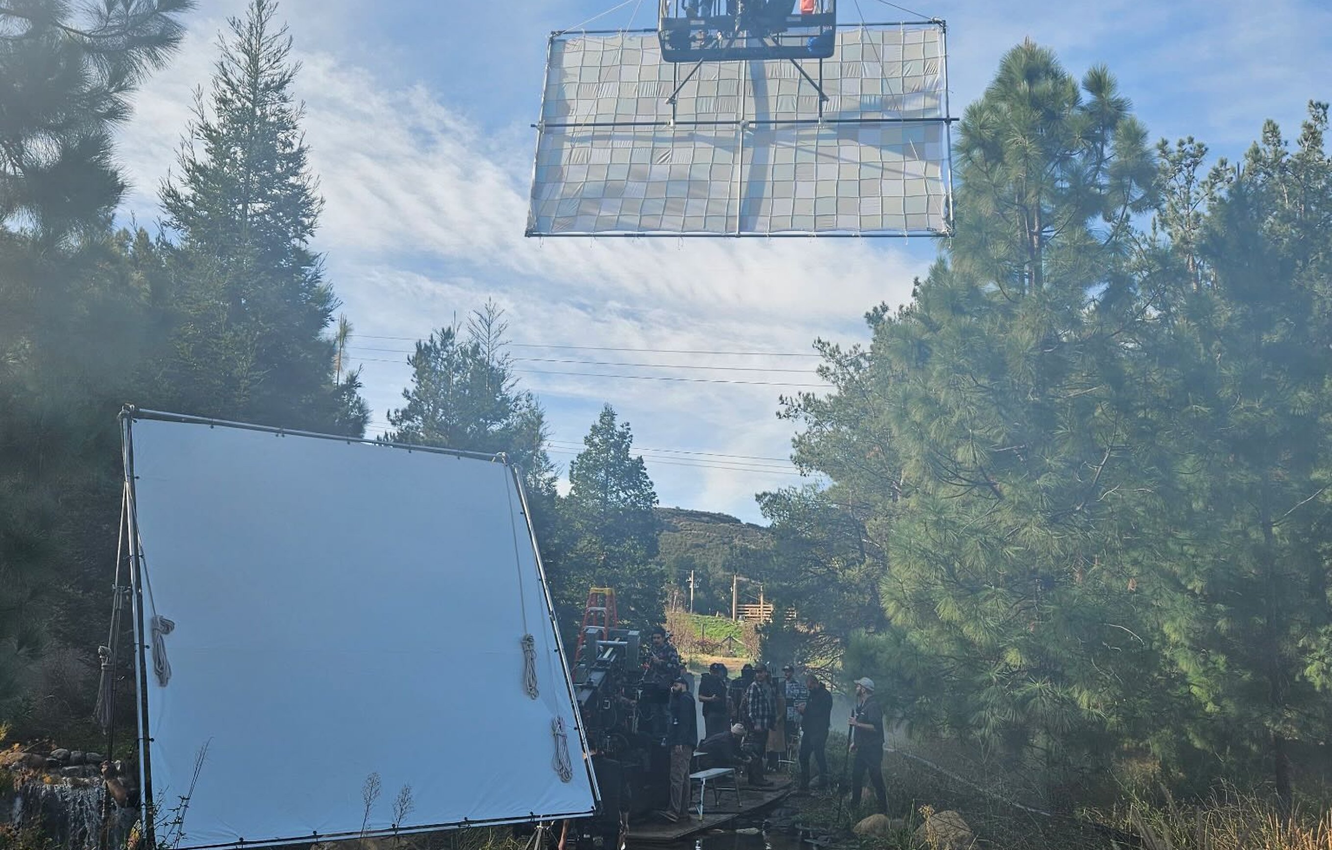 Wide view of an outdoor commercial set - Condor with 12x20 checker bounce mounted overhead above a creek lined with pines, white solid on c-stands at left, crew working in soft morning haze.