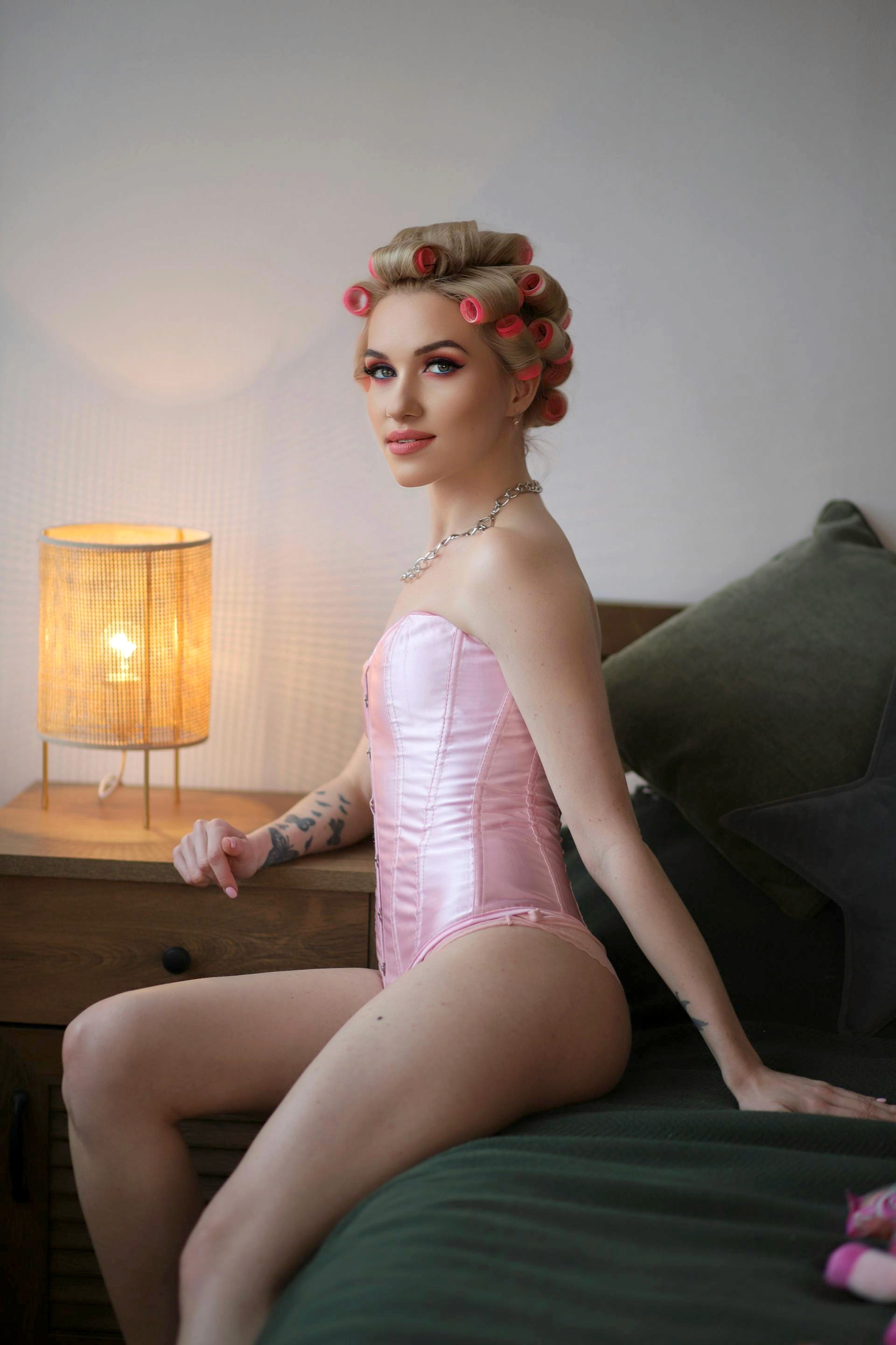A woman with blonde hair in pink rollers sitting on the edge of a bed, wearing a pink satin corset next to a glowing bedside lamp.