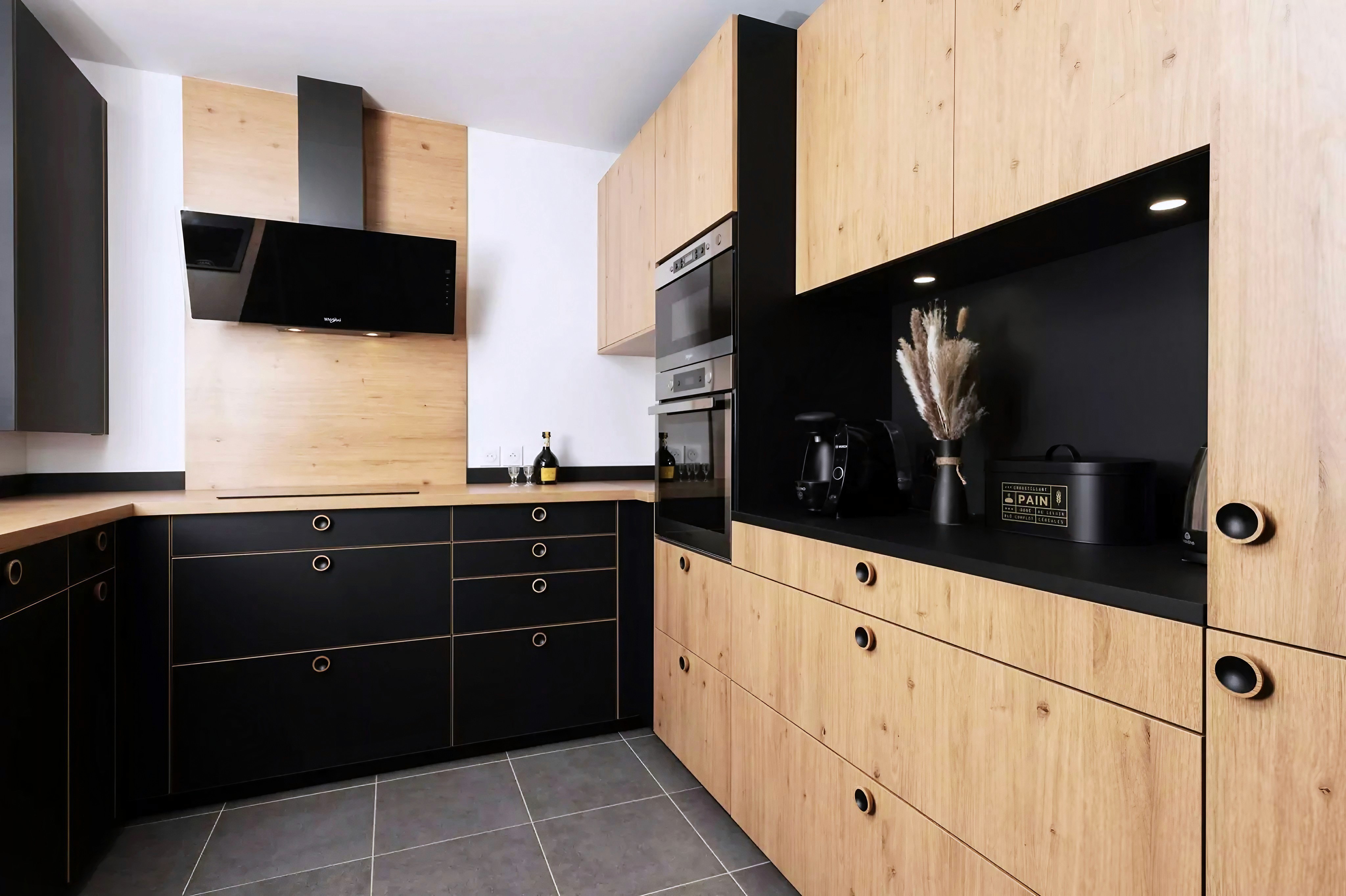 Rénovation cuisine 77 avec armoires bois noir et clair, fours encastrés, cuisinière noire et hotte assortie, sol carrelé.