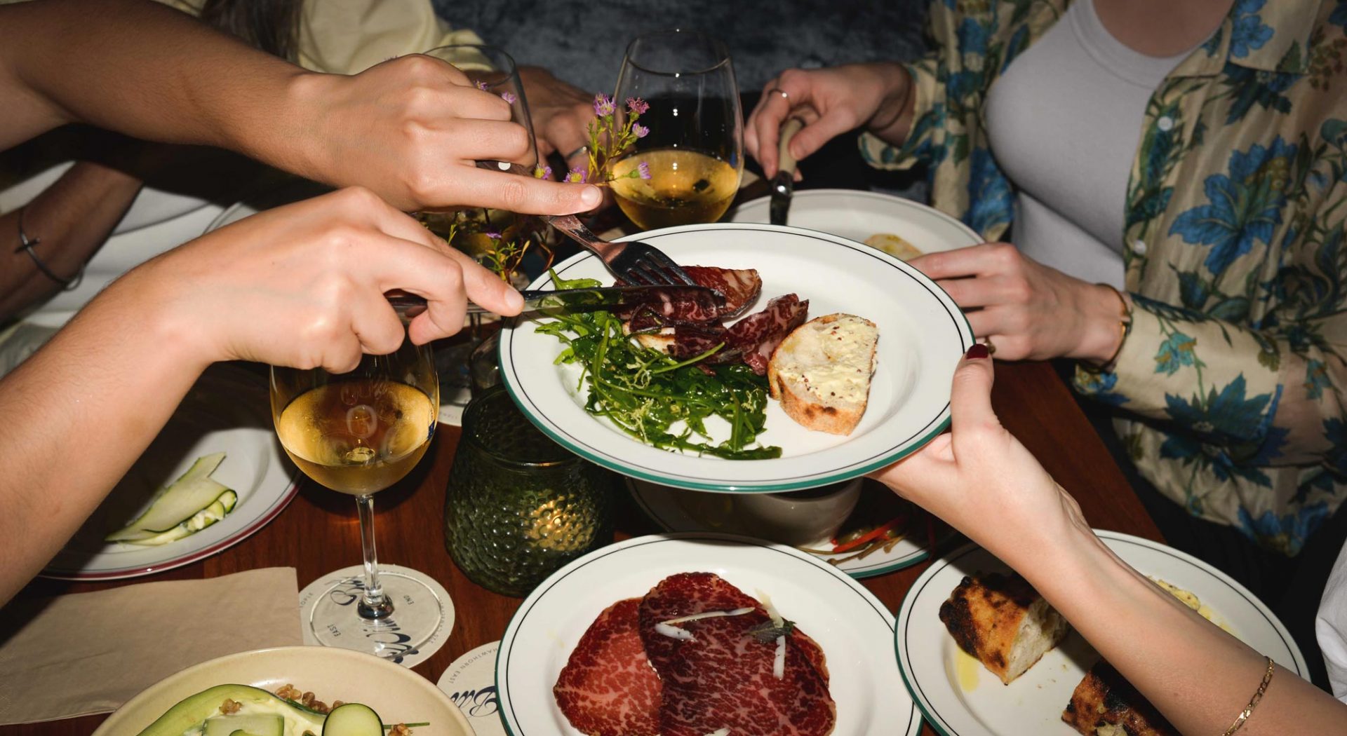 Overhead view of hands sharing a shared plate of steak and rocket, with wine glasses and cured meat plates around a restaurant table
