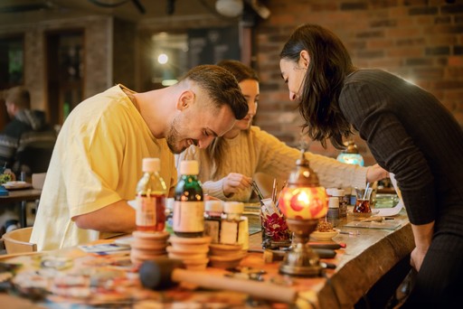 A person crafting a Turkish mosaic lamp, carefully applying multicolored beads to the ornament. The background shows a workshop setting with tools hanging on the wall.