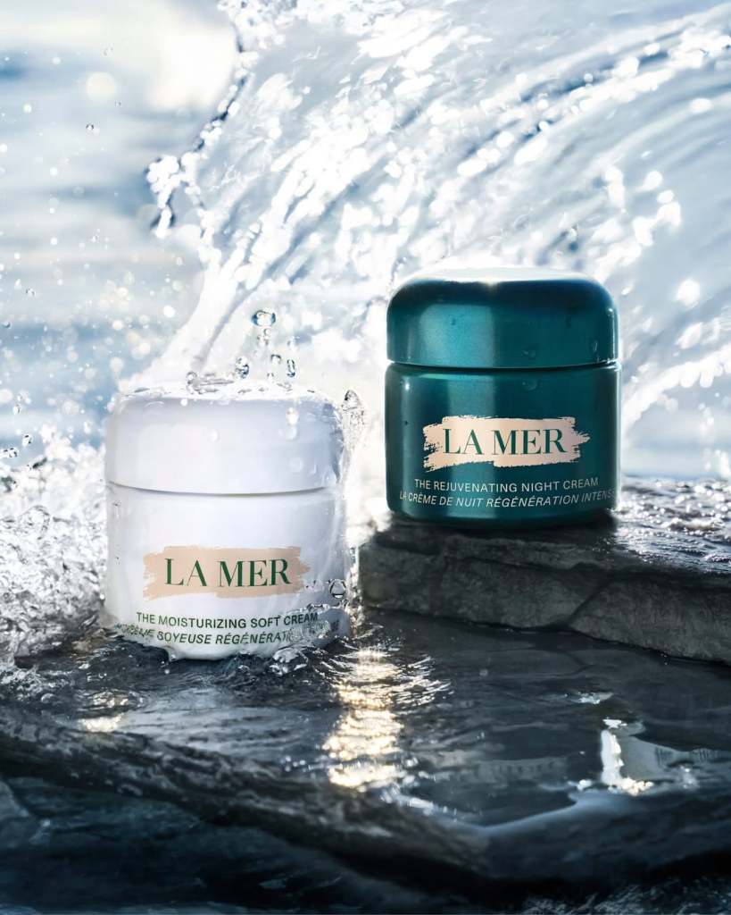 Two La Mer skincare products: a white jar of Moisturizing Soft Cream and a green jar of Rejuvenating Night Cream, positioned on a wet stone surface with splashing water in the background.