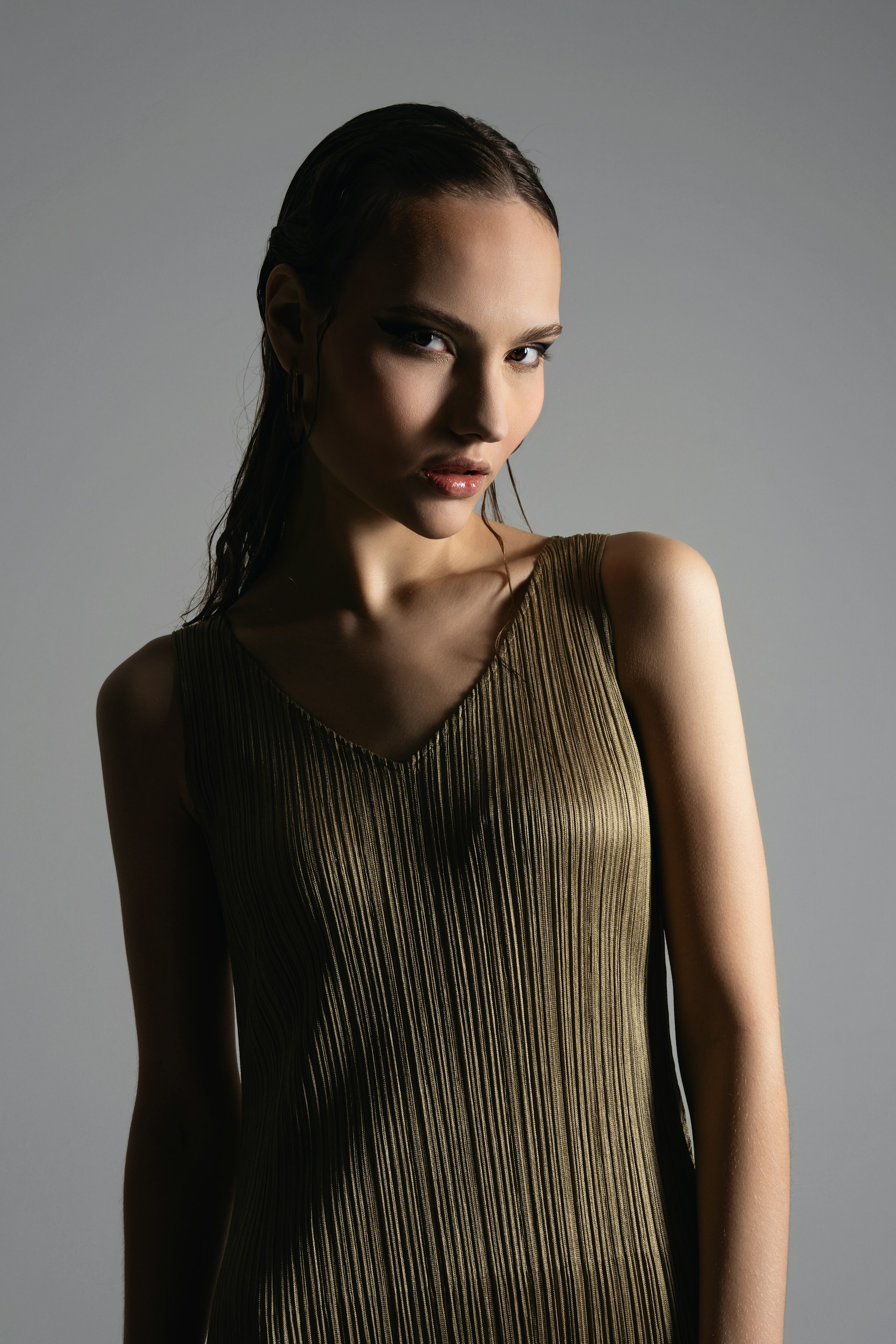 A close-up portrait of the woman with wet-look hair and bold, dark eyeliner. She is wearing the pleated green dress and posing with a slight tilt of her head under dramatic studio lighting.