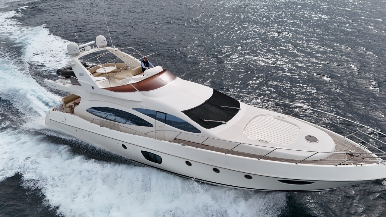Classic starboard three-quarter aerial running shot of the Azimut 68 Evolution luxury motor yacht at speed on sparkling open sea, revealing the full profile including the wraparound flybridge with cream leather seating, mahogany hardtop, side-entry door, and expansive hull windows