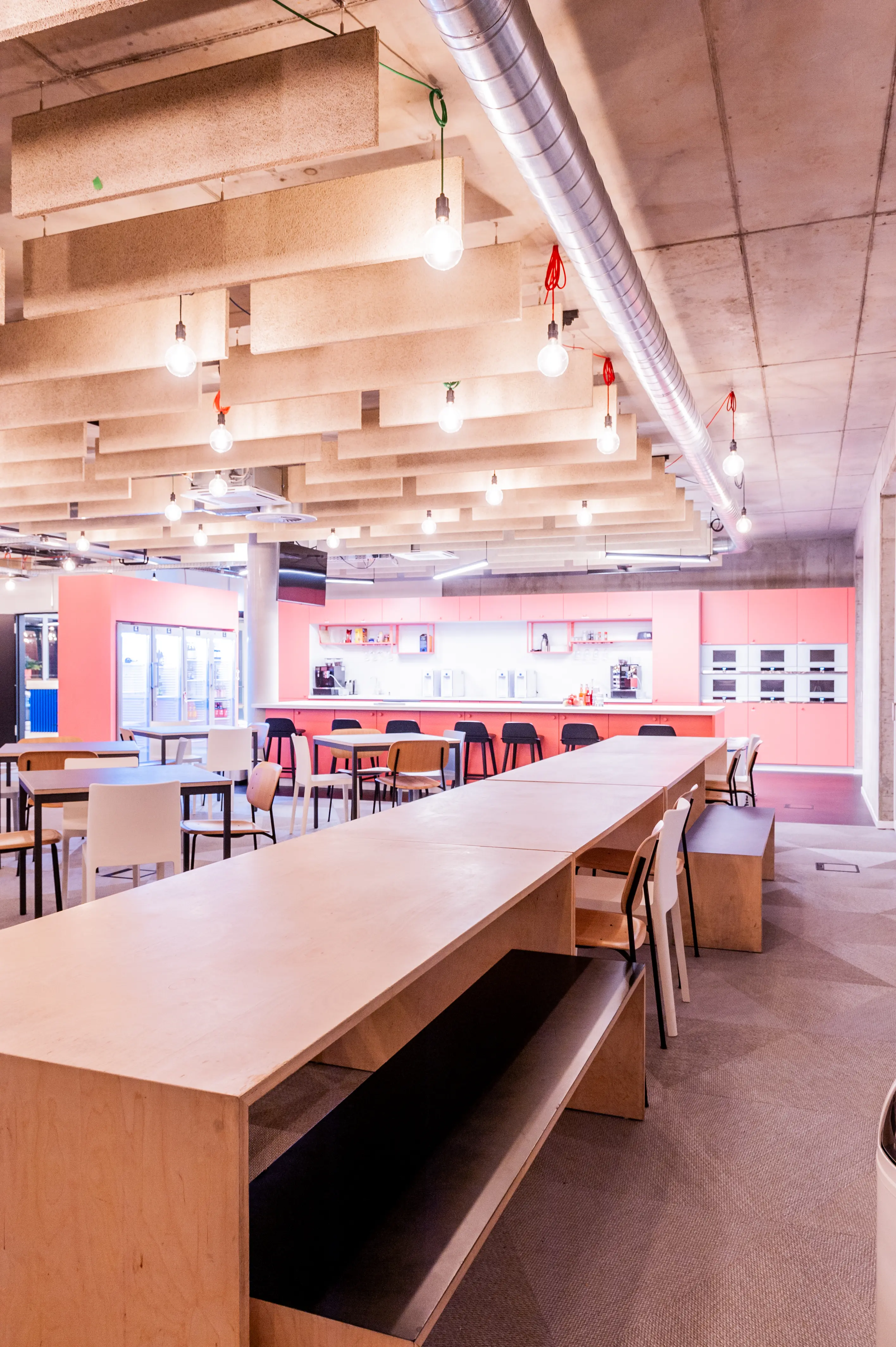 Canteen at OMIO with long plywood tables and benches, a coral-pink kitchen counter with black bar stools, bare filament bulbs and wood-fibre acoustic baffles under the concrete ceiling.