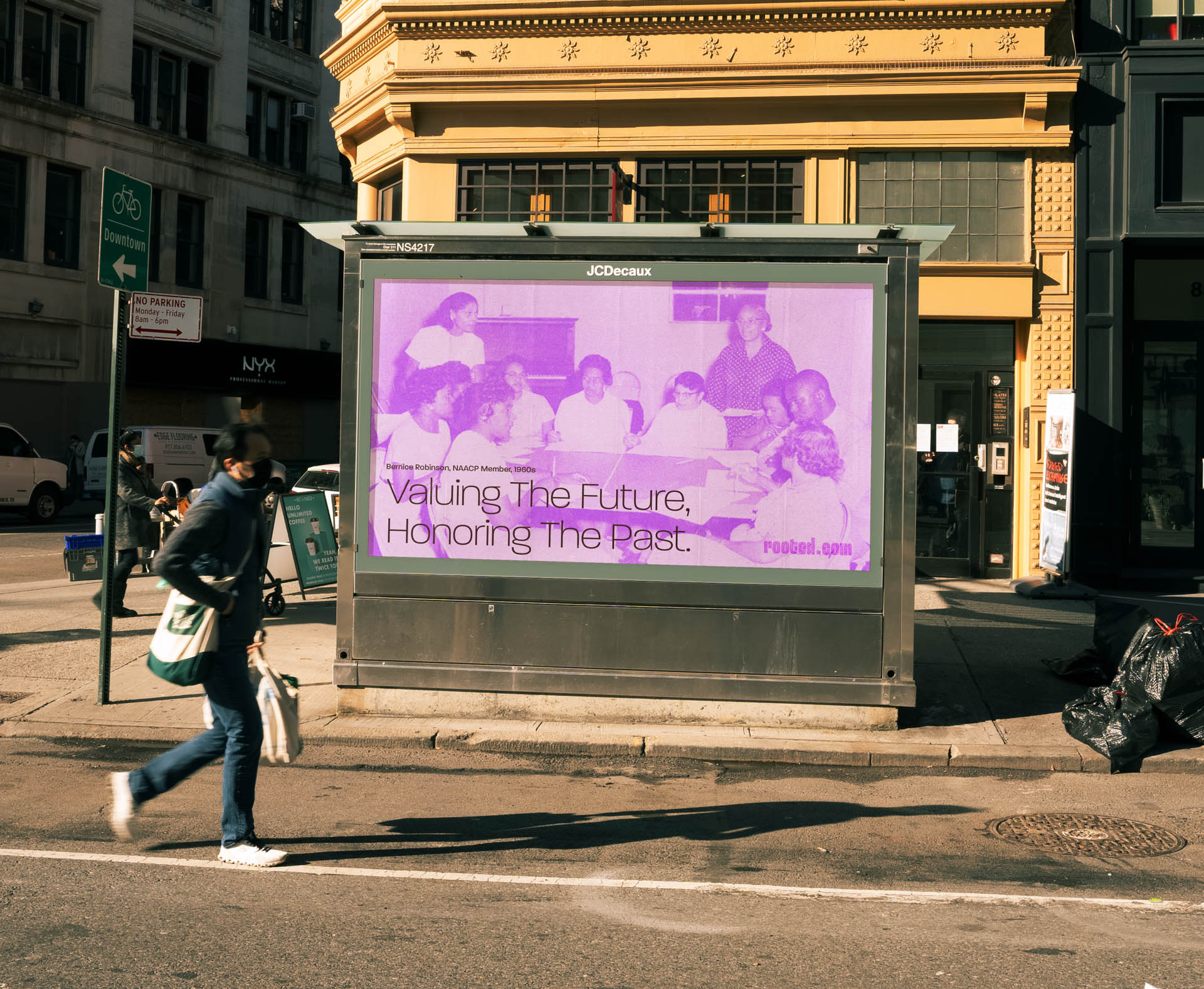 A person walks past a digital billboard displaying a purple image of multiple individuals seated at a table. The text on the billboard reads “Valuing The Future, Honoring The Past.” The billboard is mounted on a metal structure and is situated on a city sidewalk near a street with a crosswalk. Buildings are visible in the background, and a street sign is present to the left of the billboard.