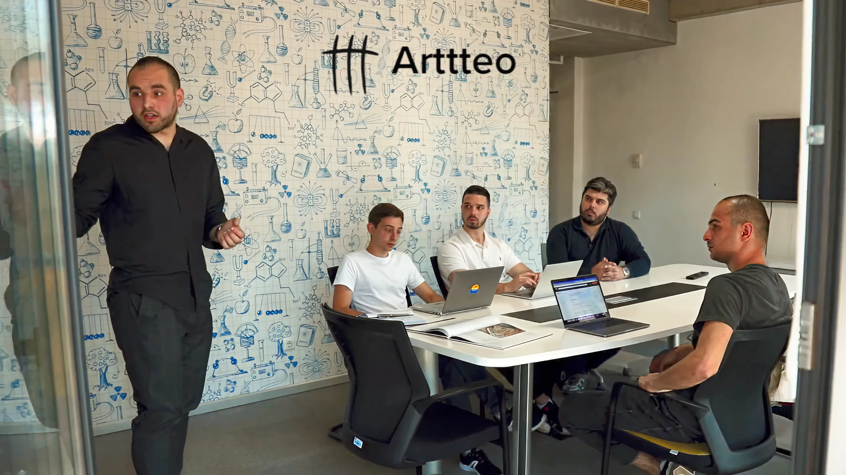Arttteo smart glasses development studio office photo showing a team meeting with five developers around a conference table against a wall featuring the Arttteo logo and illustrated laboratory mural