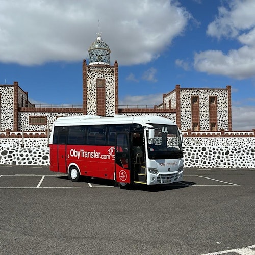 一辆标有“ObyTransfer.com”字样的红白巴士停在一栋带有石质外墙和塔楼的建筑前的空地上。