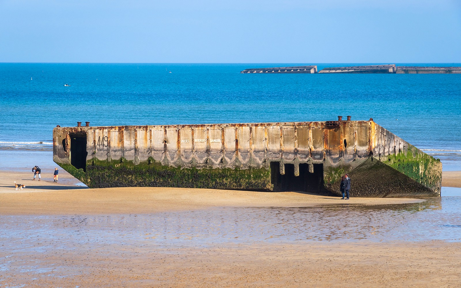 Remains of a D-Day landing craft on the beach in Caen, France, with visitors exploring nearby.
