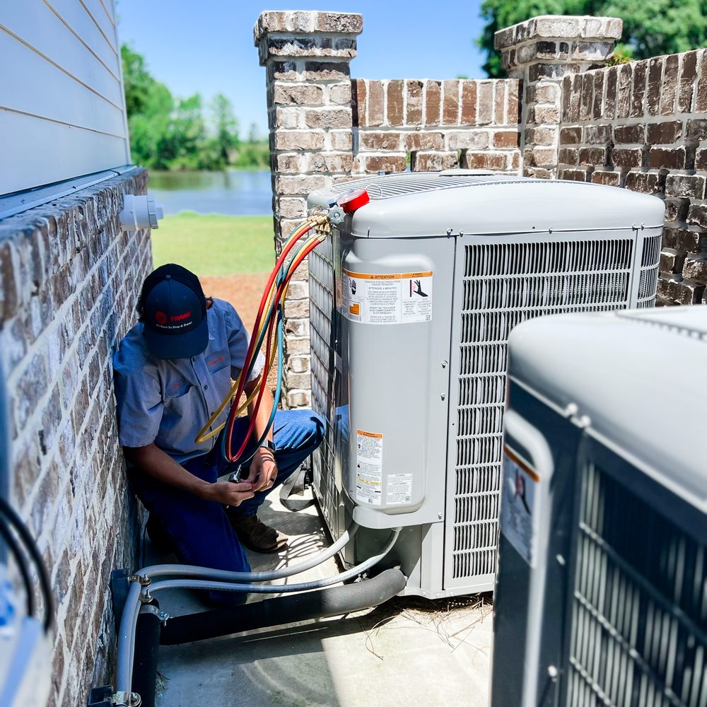 A technician repairs an outdoor air conditioning unit next to a brick wall with a lake and trees visible in the background.