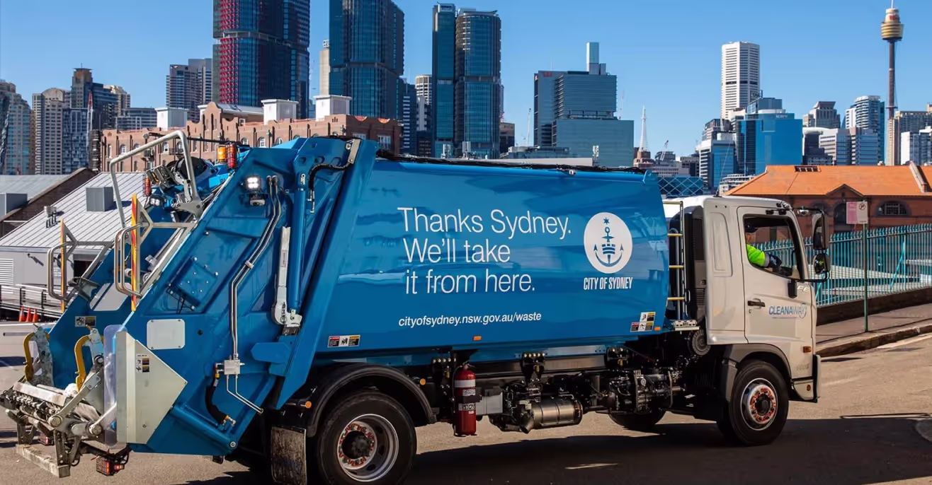 A blue City of Sydney waste collection truck lettered Thanks Sydney, we'll take it from here, parked with the city skyline behind