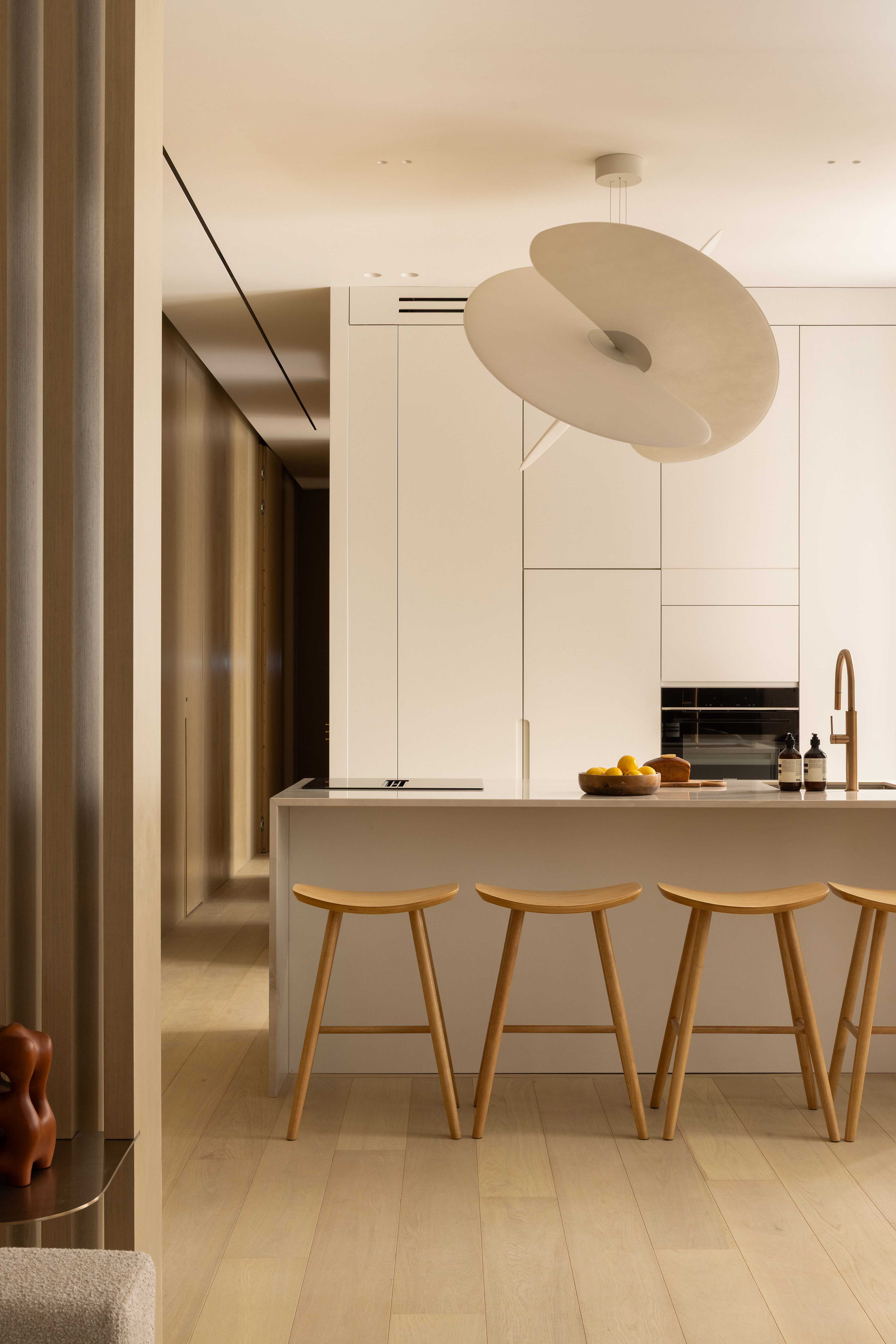 Cuisine avec îlot central, quatre tabourets en bois, suspension blanche et four encastré