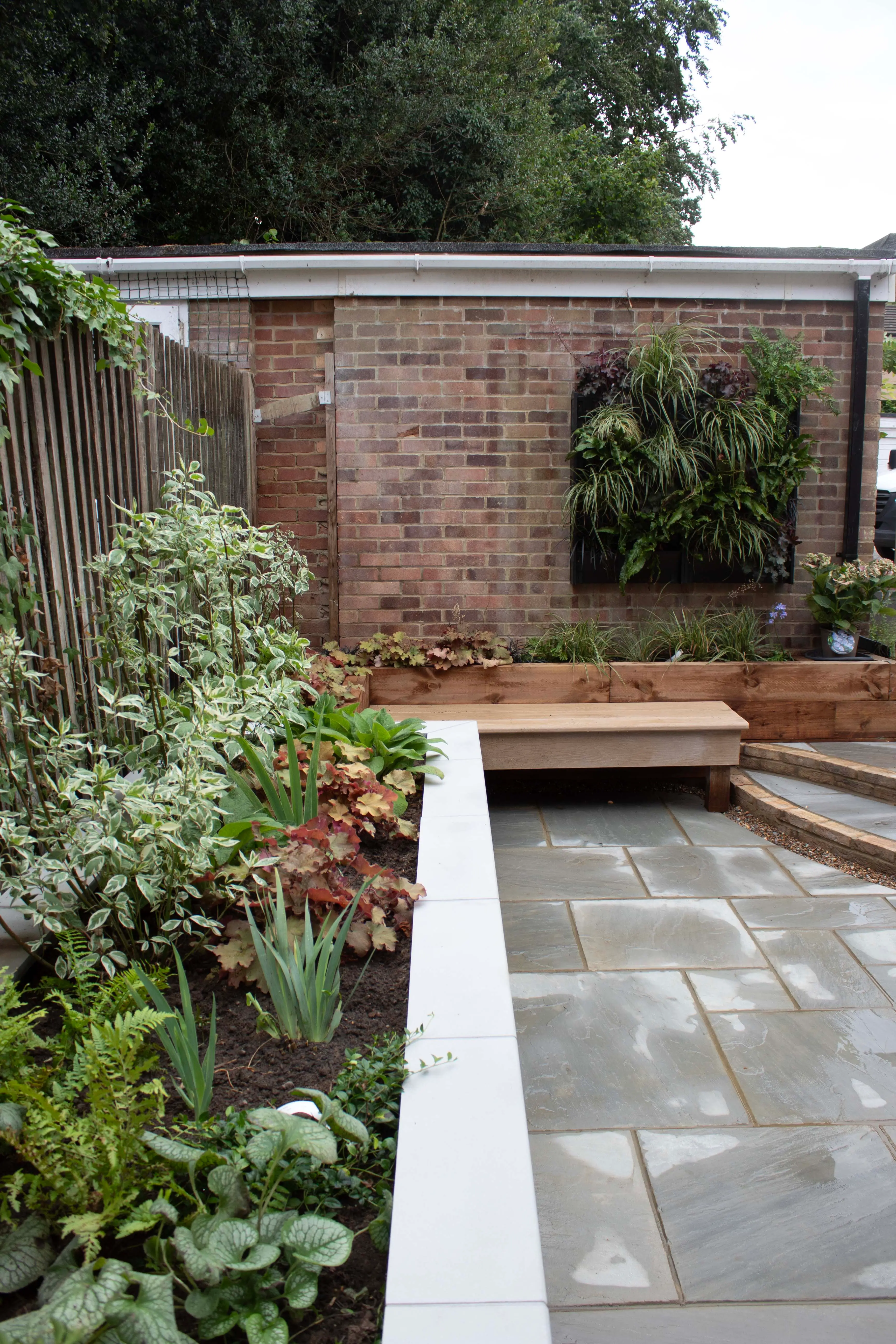 A serene garden path leads to a textured brick wall, surrounded by lush greenery and plants.