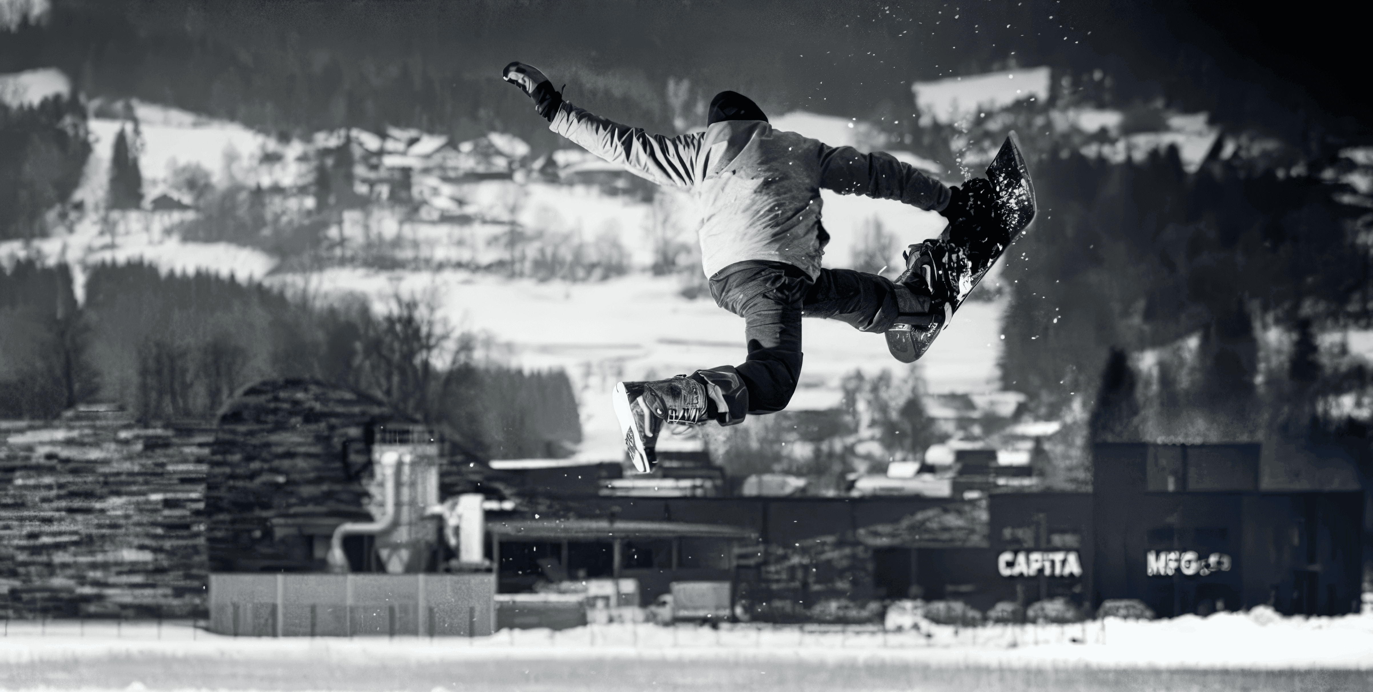 MW Interior - Snowboarder bei Sprung in einem Snowpark vor alpiner Winterkulisse in Schwarz-Weiß" Beschreibung: Dynamische Atmosphäre eines Snowpark-Standorts im Alpenraum – ein Referenzprojekt, bei dem Einrichtungs- und Möbellösungen für den Freizeit- und Gastrobereich realisiert wurden.